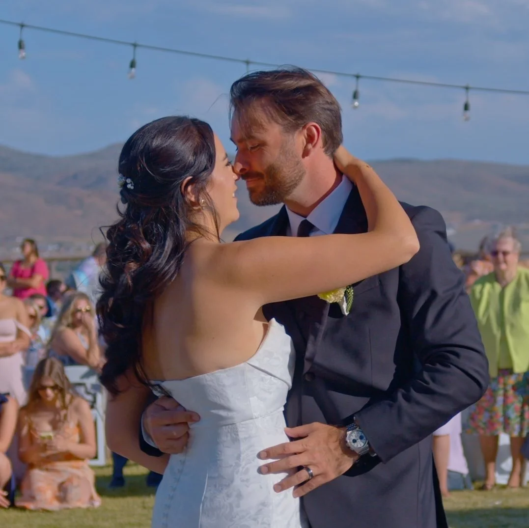 A bride and groom share a romantic dance outdoors at sunset, surrounded by guests, with mountains in the background.