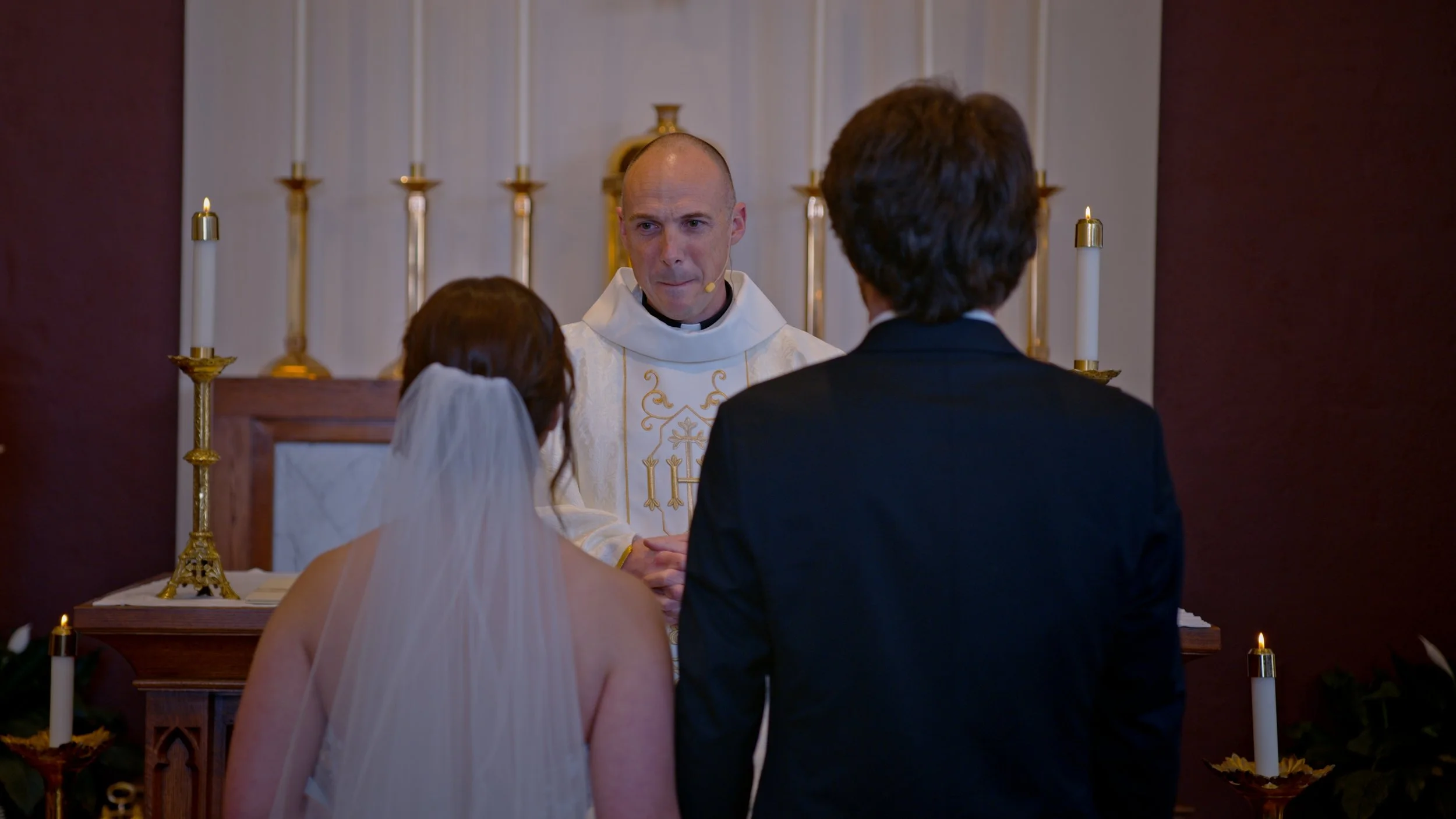A wedding ceremony with a priest officiating at the altar, a bride in a white veil and gown, and a groom in a black tuxedo holding hands.