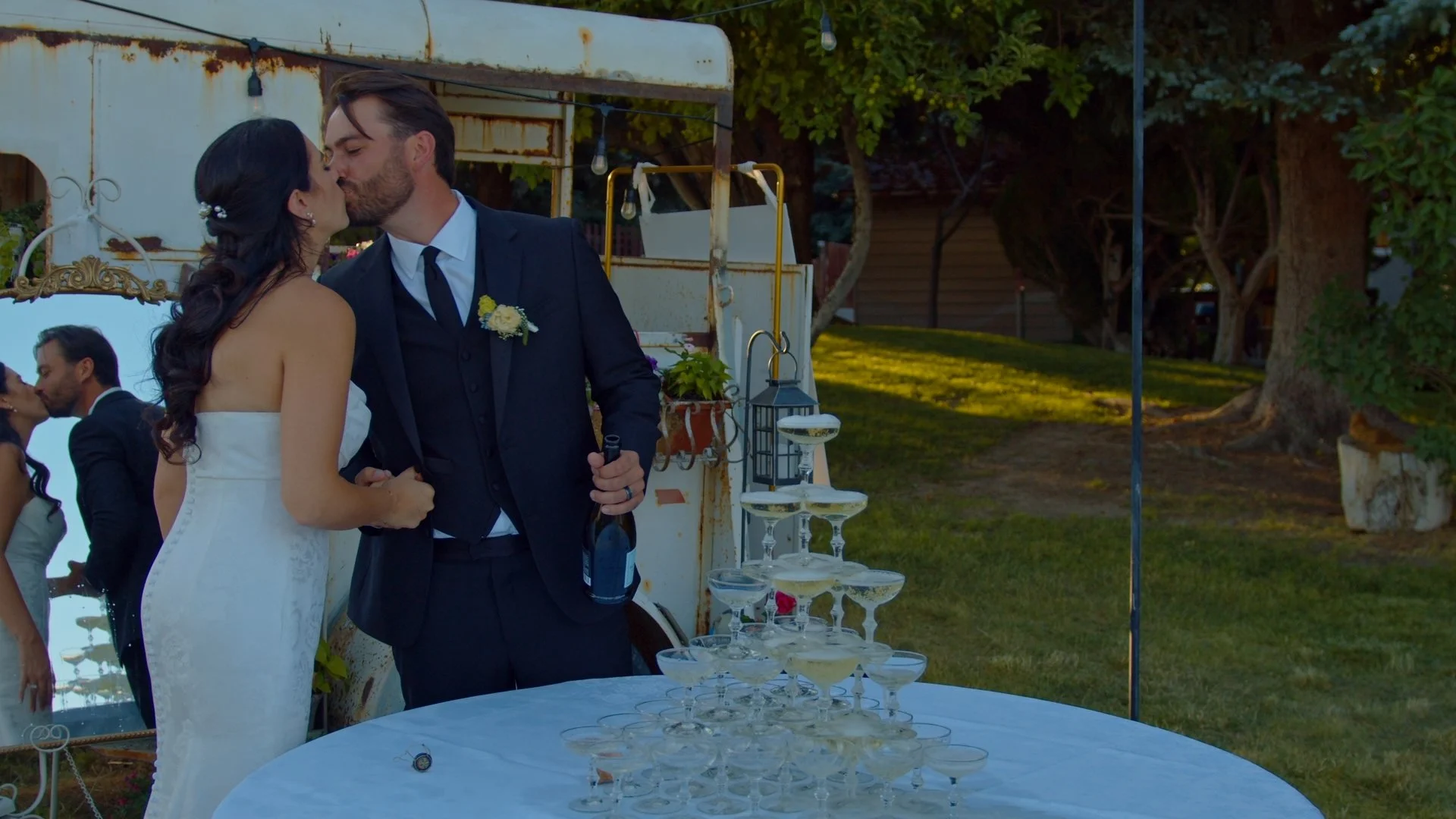 A bride and groom kissing outdoors near a champagne tower at a wedding reception, with guests in the background and trees in the distance.