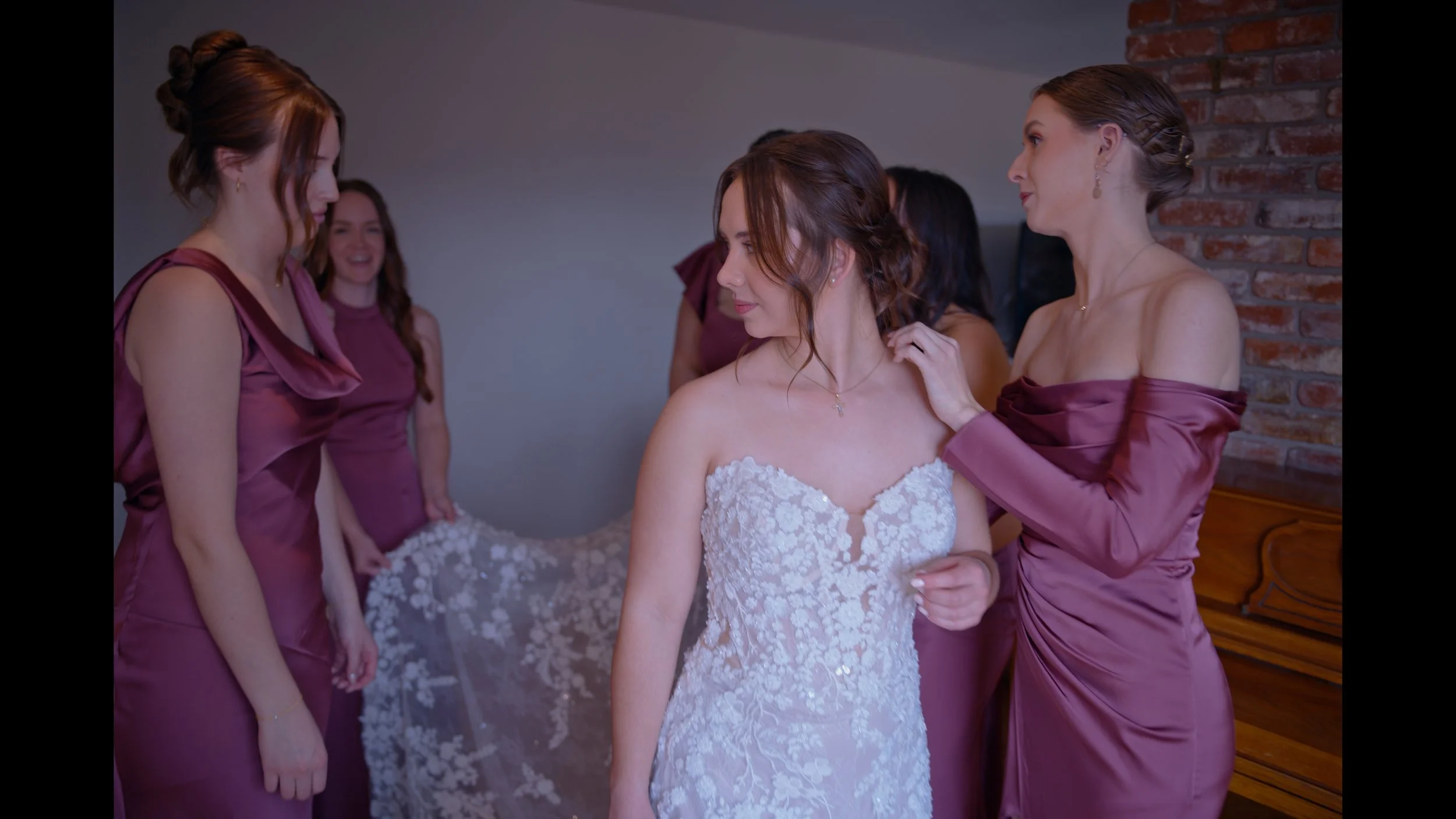 A bride in a white lace wedding dress surrounded by four bridesmaids wearing matching purple satin dresses in a room with a brick wall.