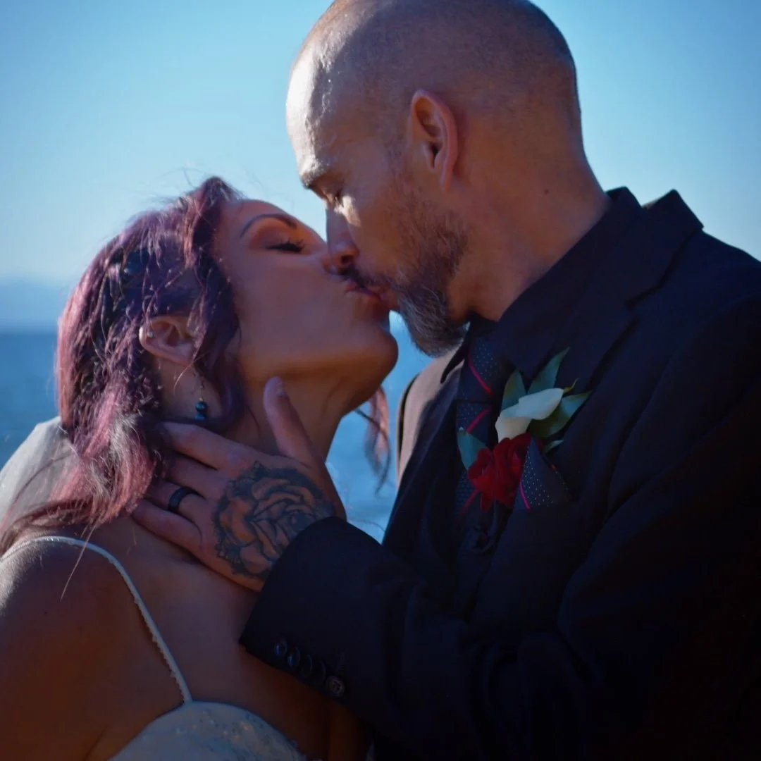 A man and woman kissing outdoors, with the man wearing a dark suit and the woman in a white dress, during daytime.