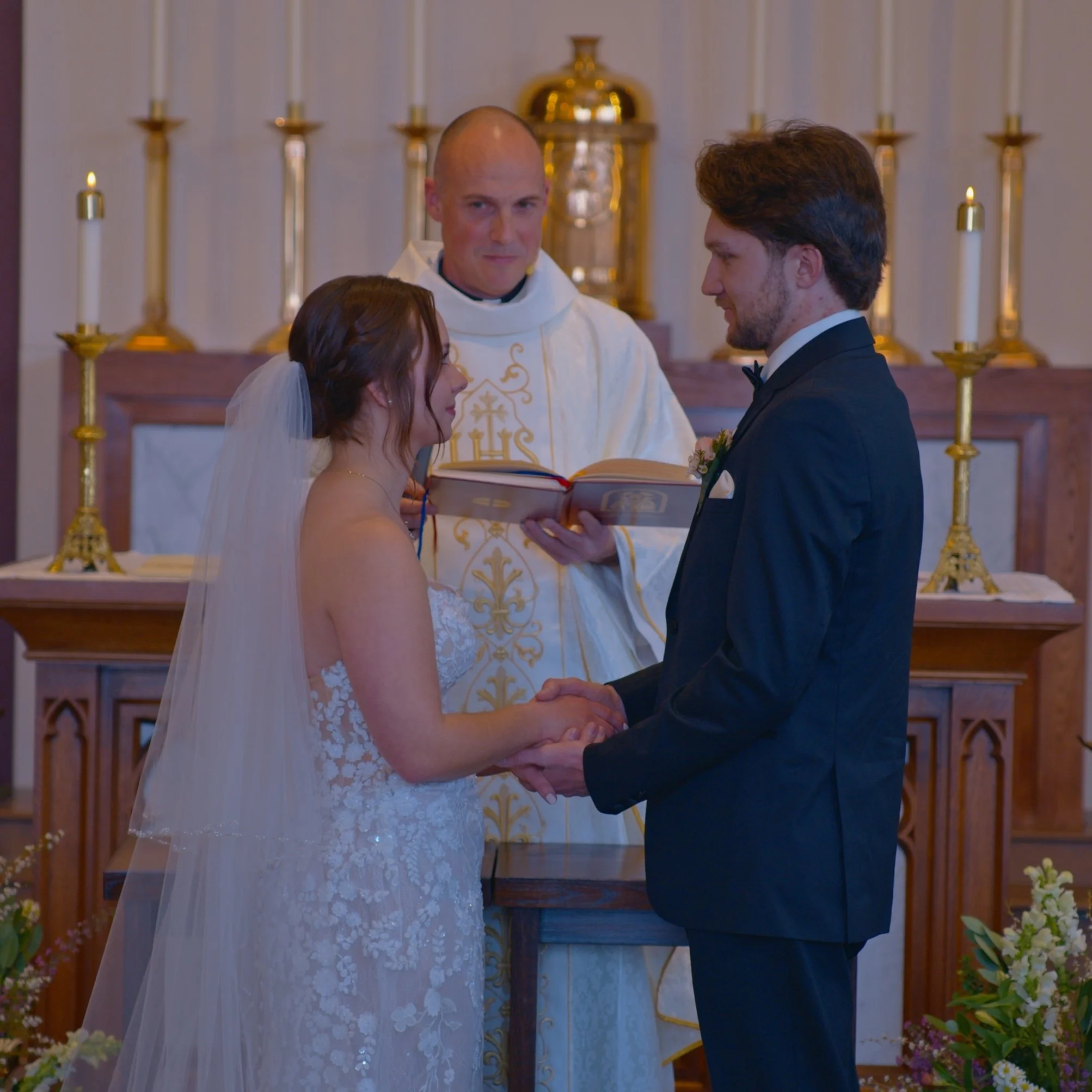 A bride and groom exchange vows in a church during their wedding ceremony, with a priest officiating, behind them are tall candles and religious artifacts.