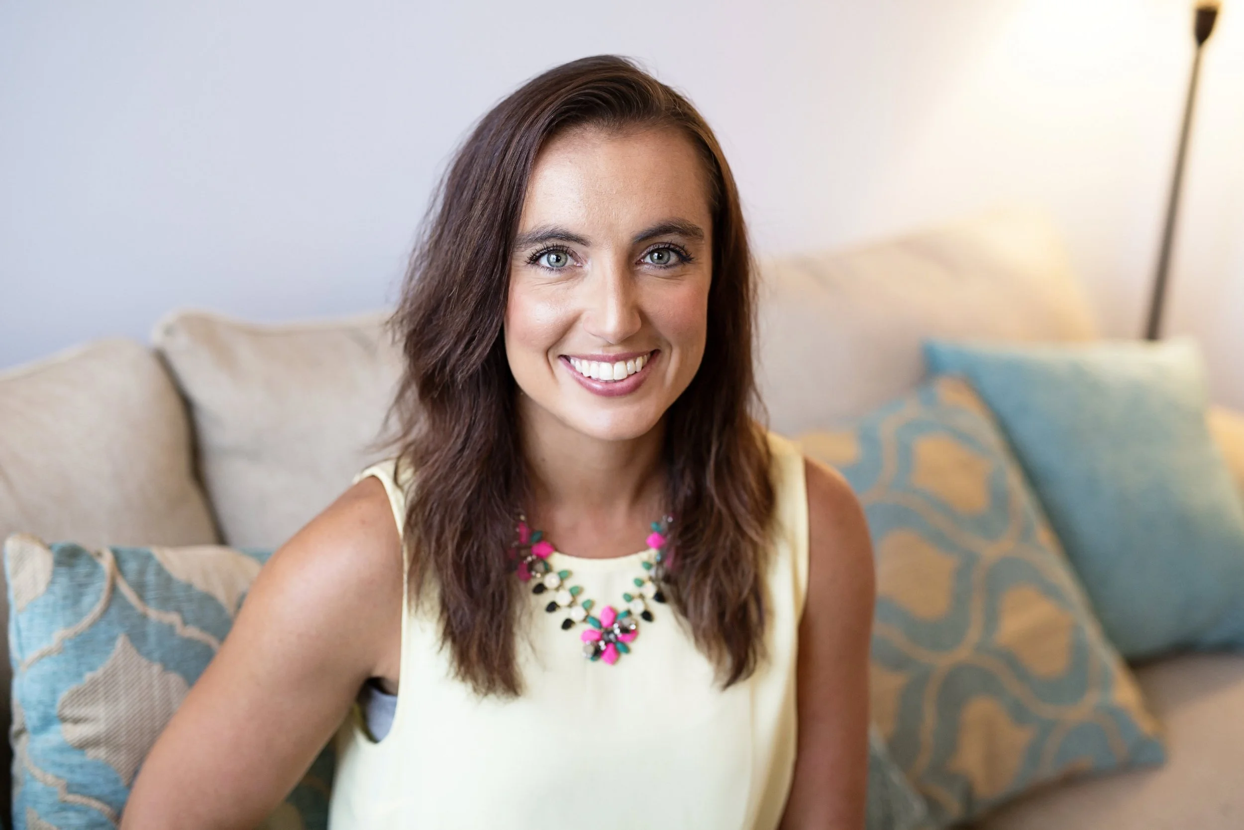 Close-up of a smiling woman with brown hair, blue eyes, wearing a yellow top and colorful beaded necklace, sitting on a patterned blue and beige sofa with a pillow behind her, in a well-lit room.
