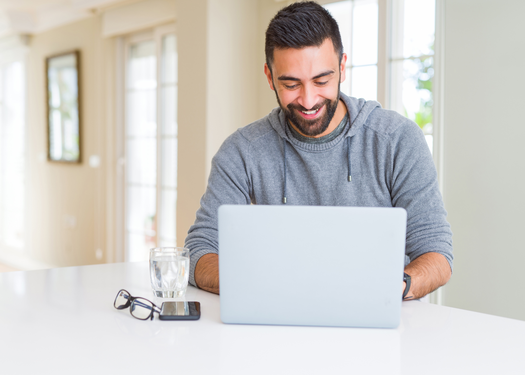 A smiling man with dark hair and a beard, wearing a gray hoodie, looking at a laptop while sitting at a white table in a bright room with large windows.