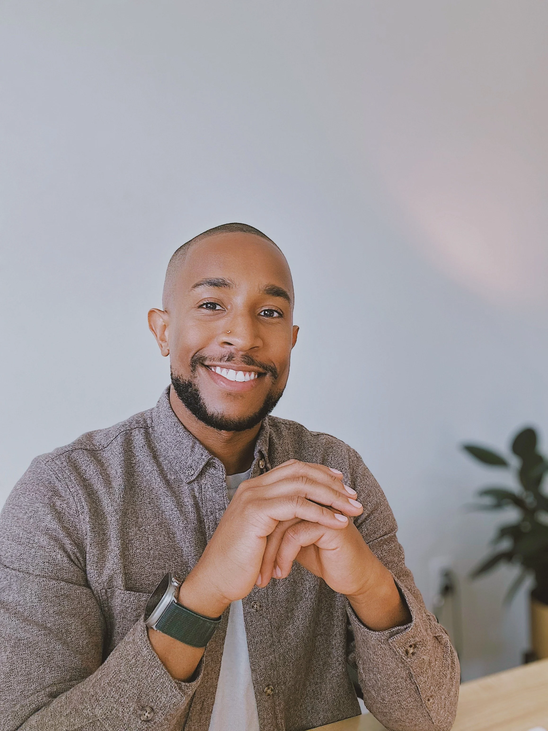 Smiling Black man in a gray button-up shirt posing with clasped hands indoors