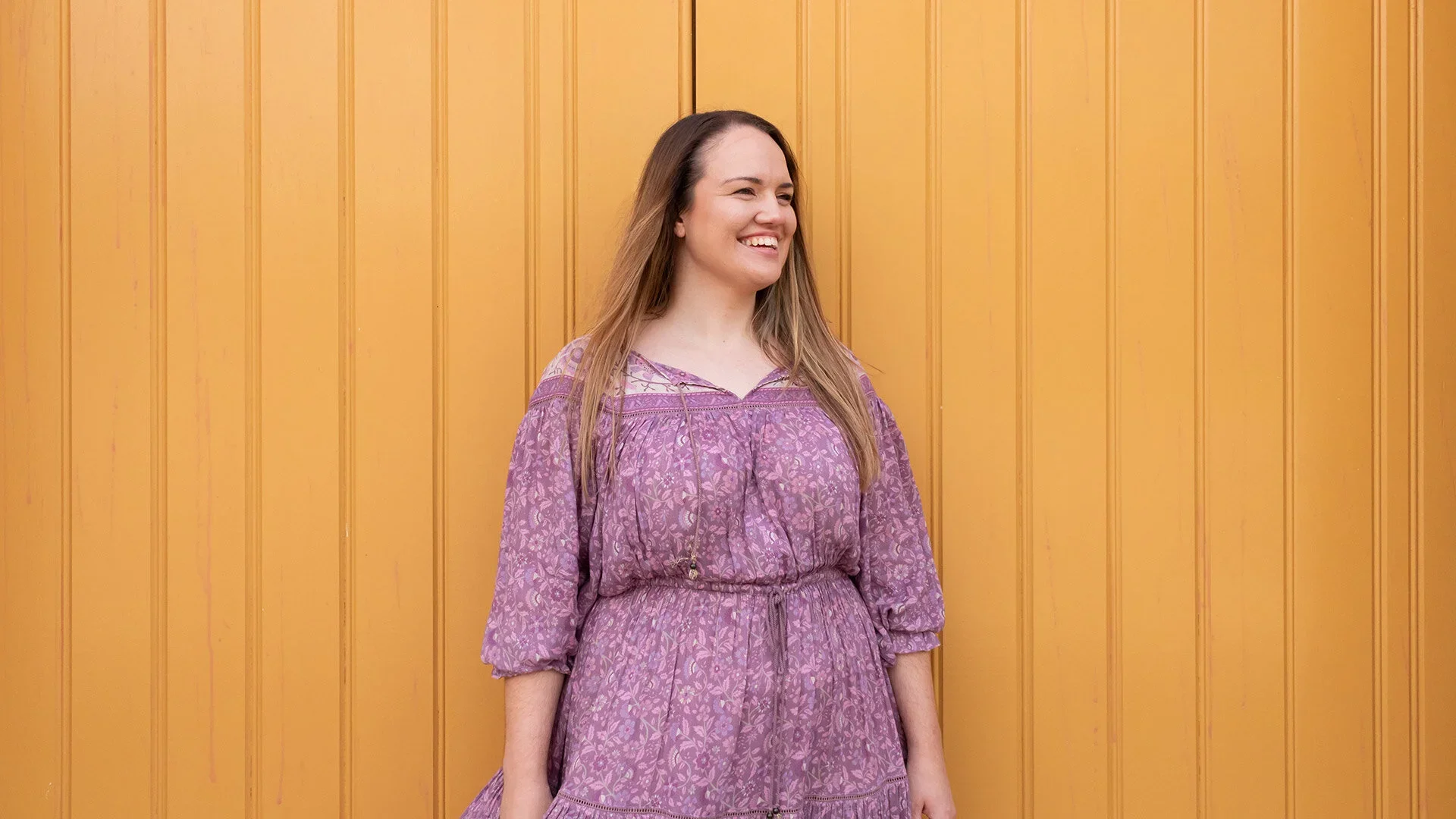 A woman with long brown hair, wearing a purple patterned dress, smiling and standing in front of a yellow wooden wall.