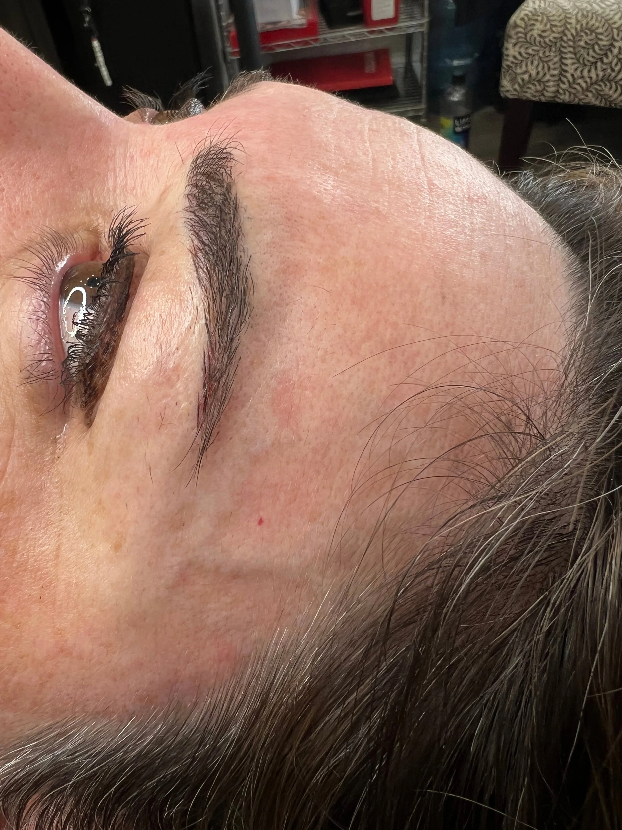 A close-up of a person's face with partially closed eyes, dark eyelashes, and brown eyebrows, lying down with a background of various objects and furniture.