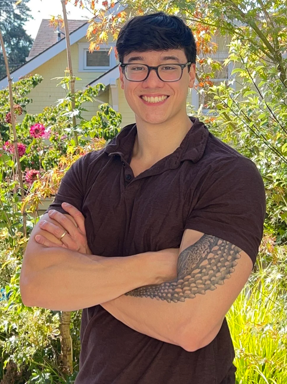 A smiling young man with glasses and a tattoo on his left arm, standing in a garden with green plants and pink flowers, and a house with a sloped roof in the background.
