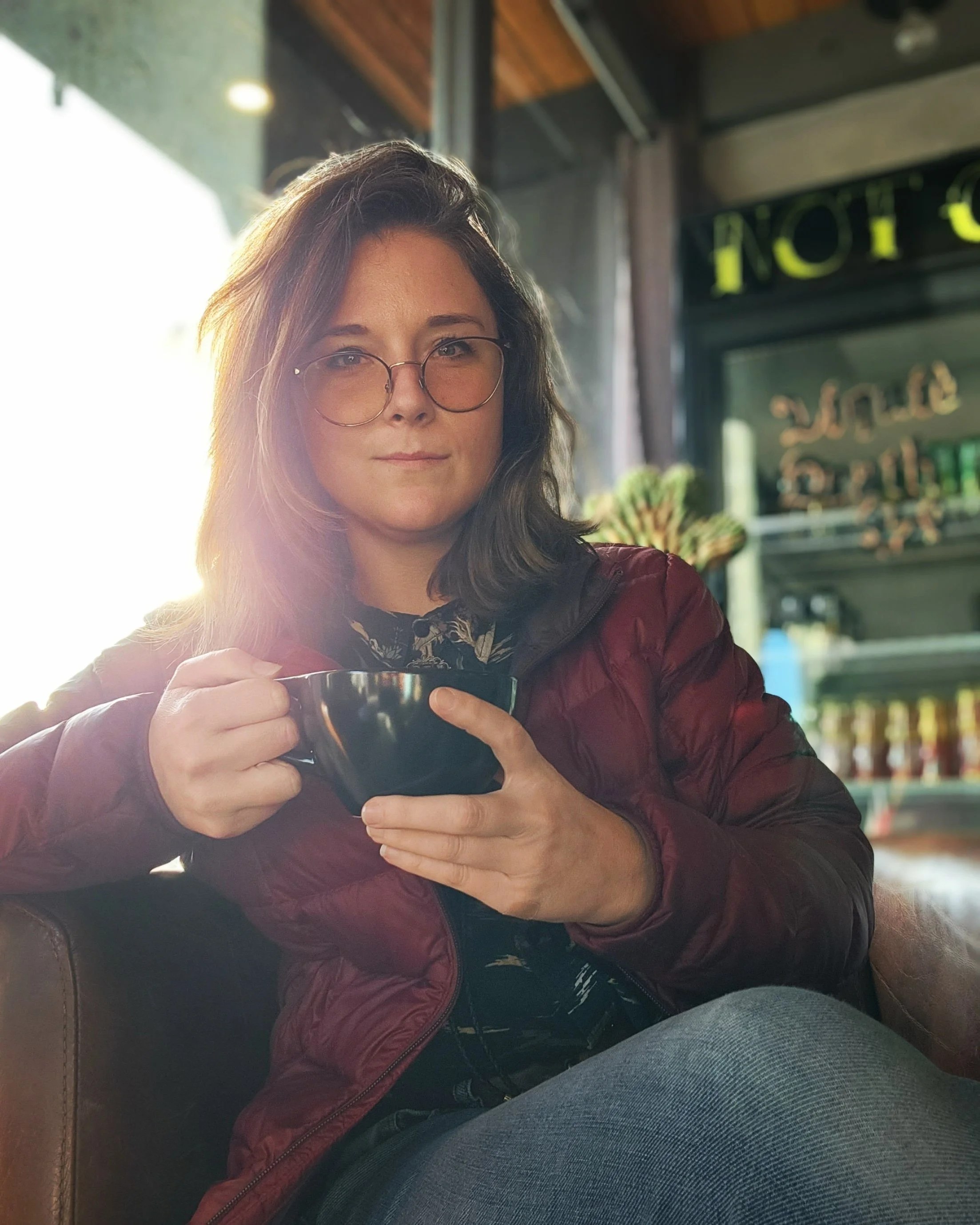 A woman with glasses, shoulder-length dark hair, and a light complexion sitting inside a cafe or restaurant, holding a dark-colored cup with both hands. She is wearing a maroon jacket and a patterned shirt. The background shows wooden ceiling beams, indoor plants, and a partially visible sign with the word 'NOT'.