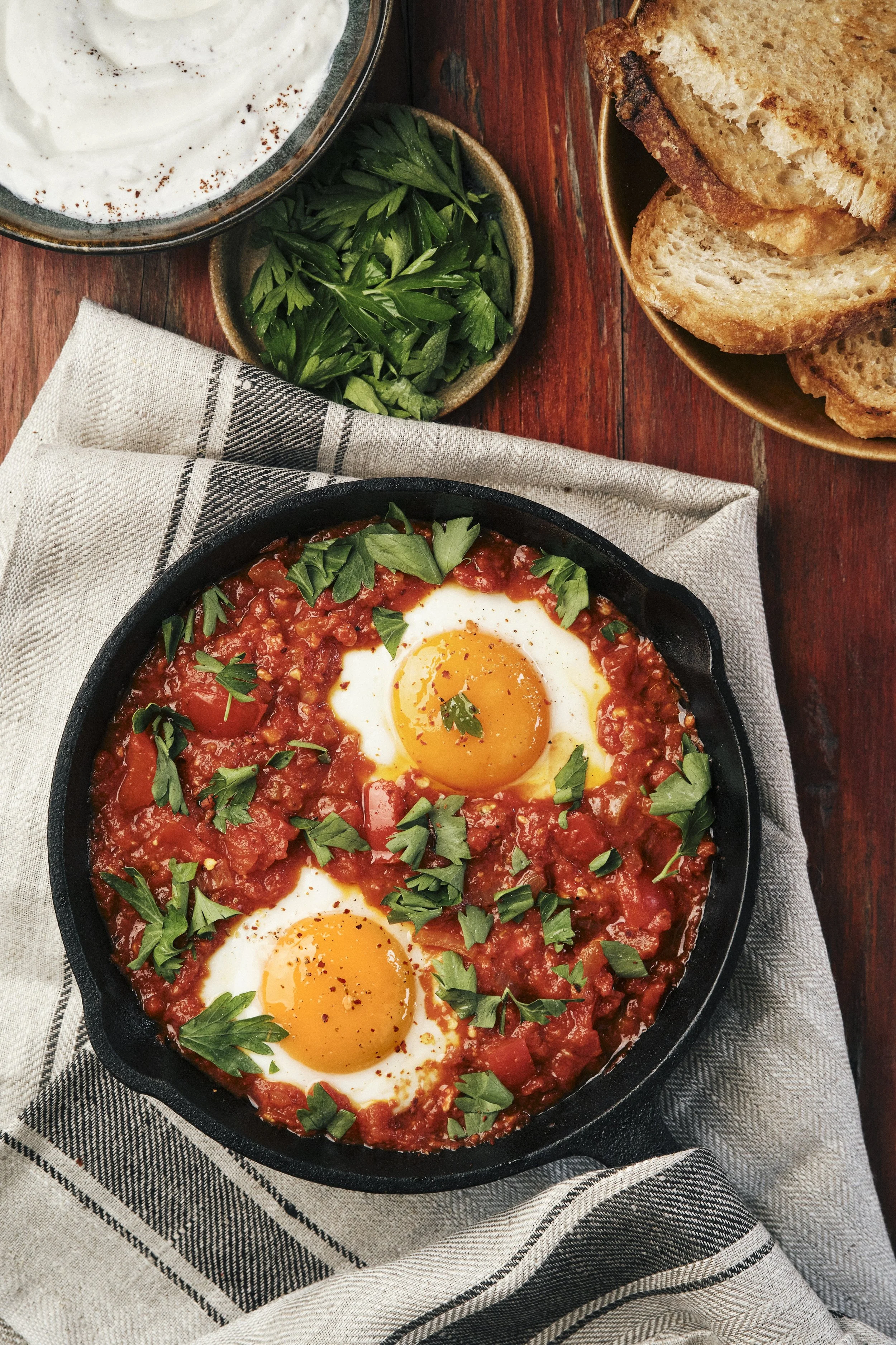Top-view food photography of shakshuka with eggs cooked in rich tomato sauce, topped with fresh parsley and served with toasted bread