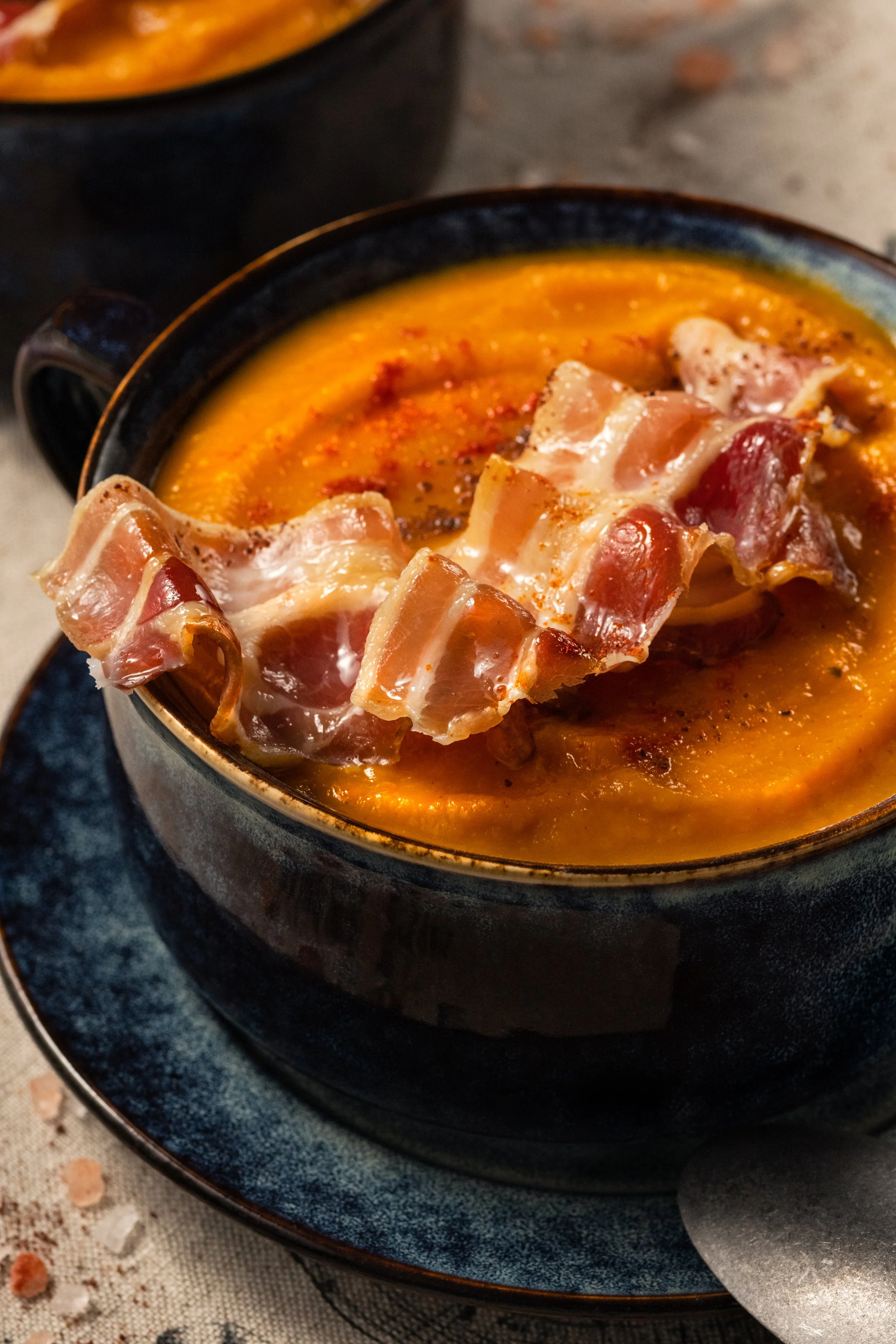 Professional food photography of creamy pumpkin soup topped with crispy prosciutto and spices, styled in a dark ceramic bowl for restaurants, food brands and editorial content