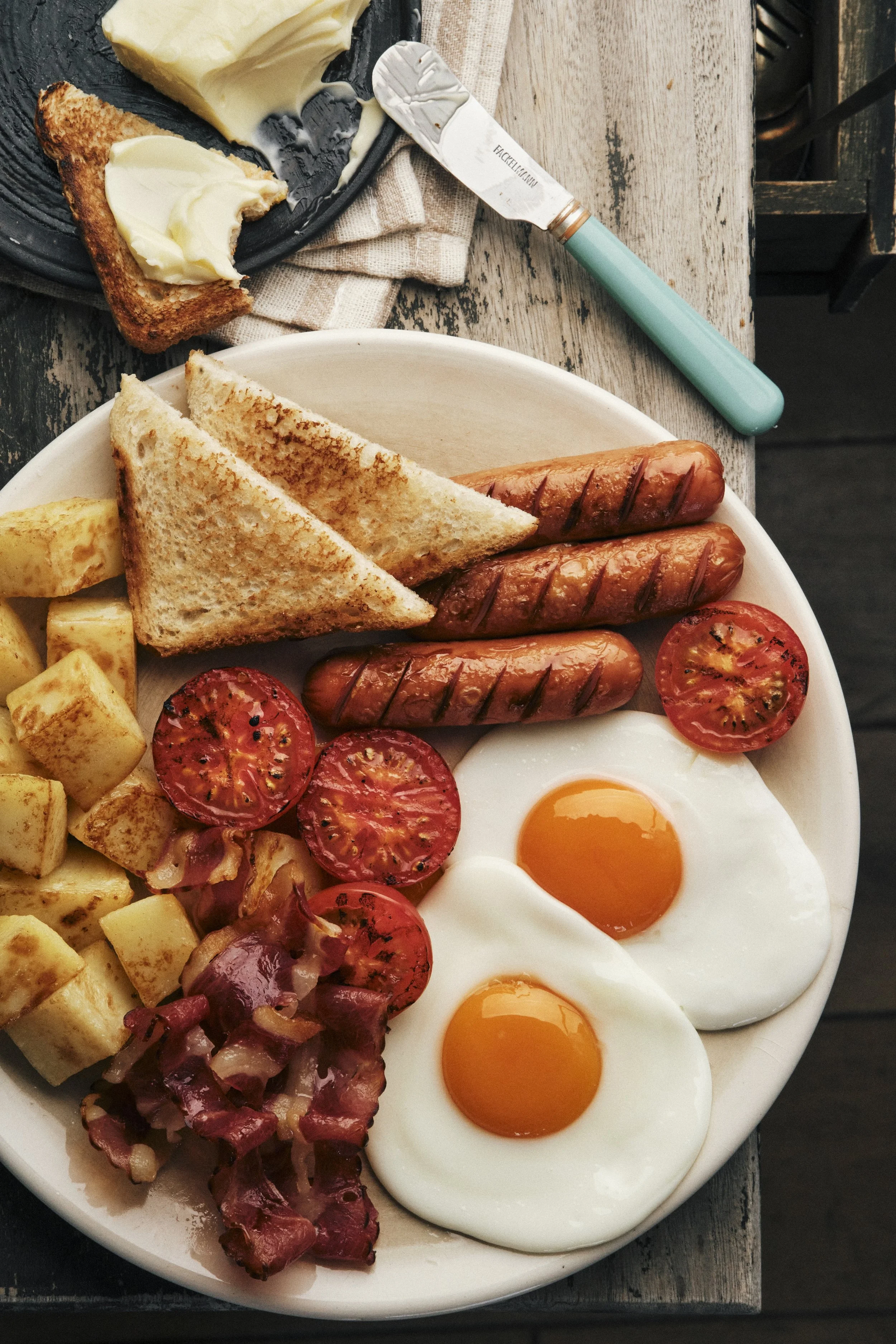 Top-view food photography of a hearty breakfast with fried eggs, grilled sausages, crispy bacon, toast, roasted potatoes and tomatoes