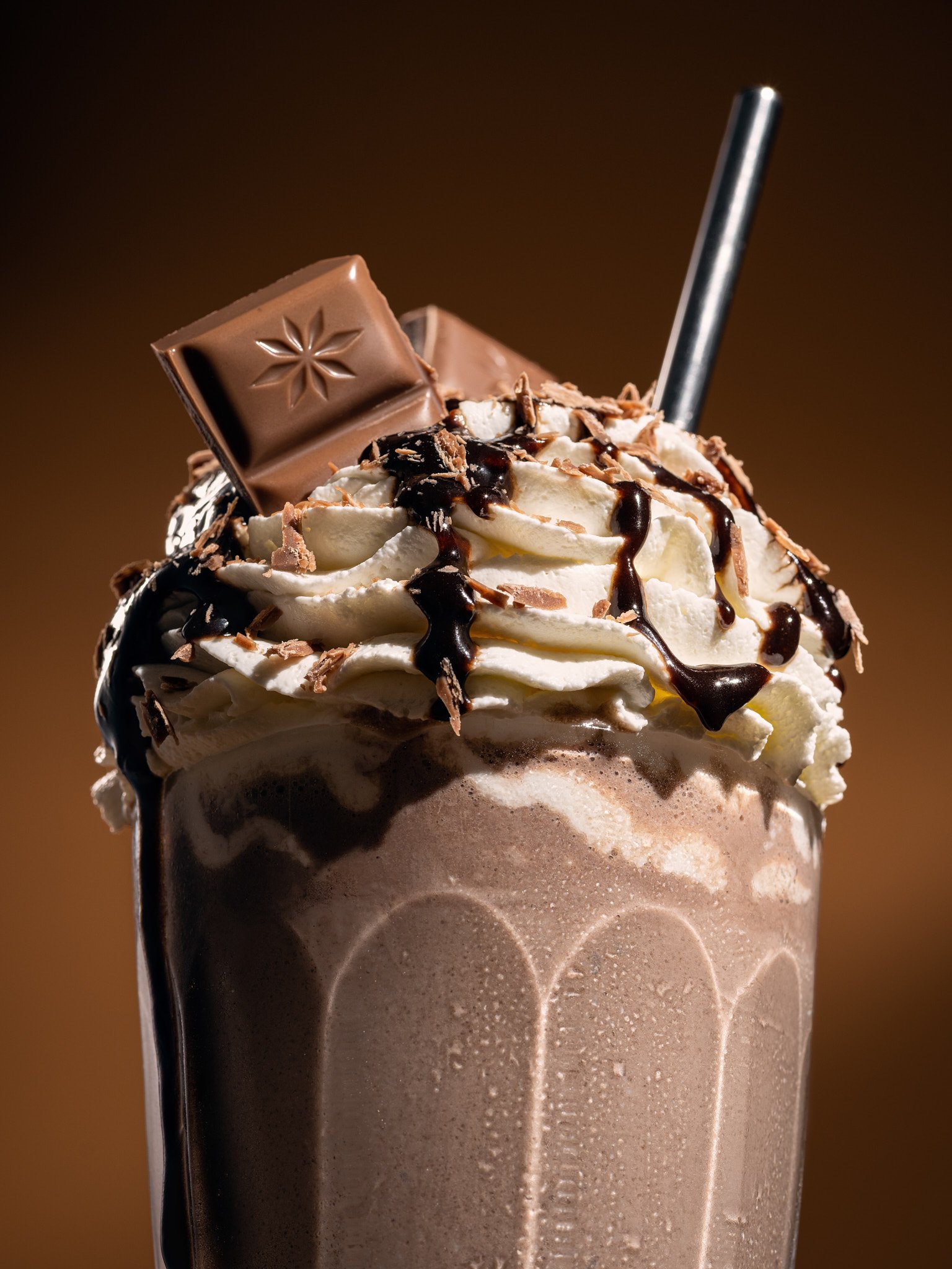 A milkshake topped with whipped cream, chocolate syrup, chocolate shavings, and a piece of chocolate with a star-shaped design, served with a straw.