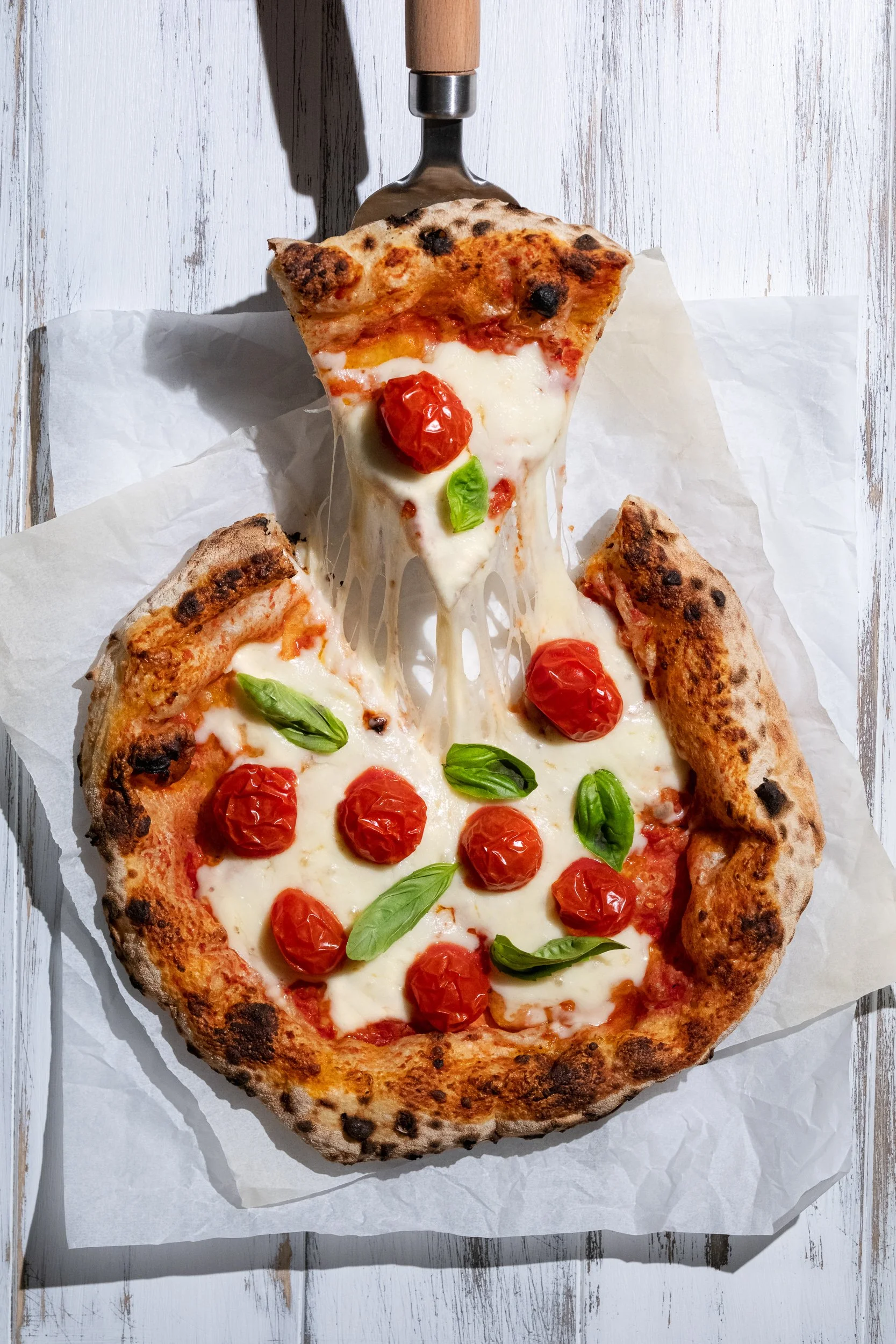 Close-up of a slice of pepperoni pizza with melted cheese, cherry tomatoes, and basil on parchment paper, on a white wooden surface.