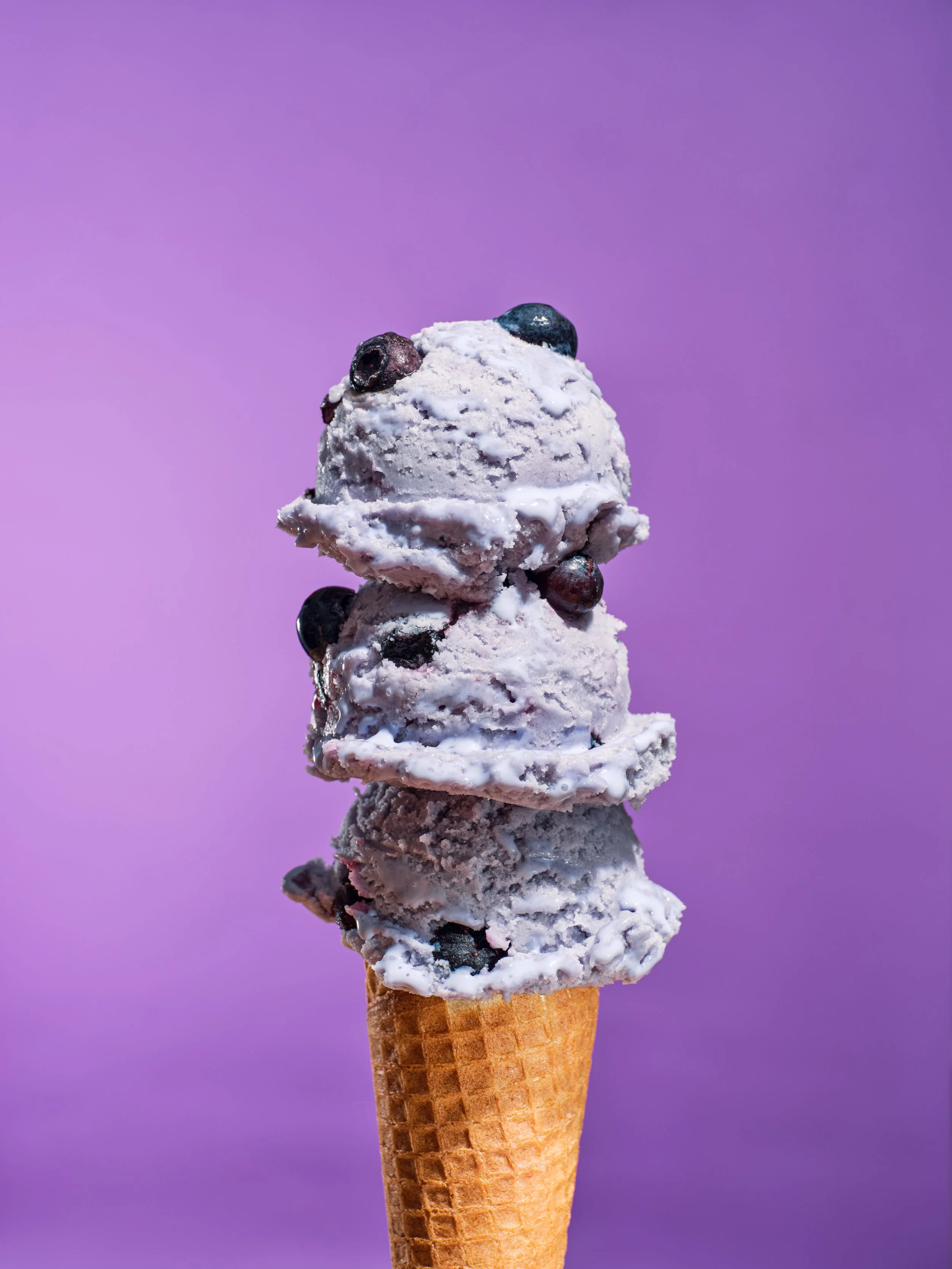 Studio food photography of three scoops of blueberry ice cream with whole blueberries in a waffle cone against a vibrant purple background