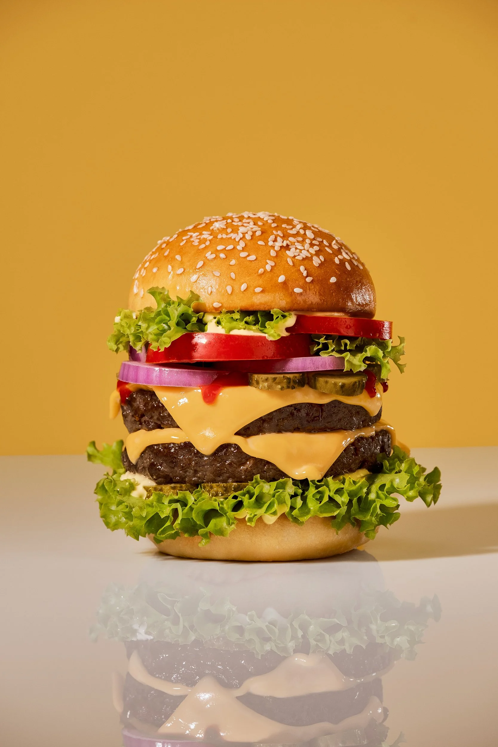 Studio food photography of a double cheeseburger with melted cheese, beef patties, lettuce, tomato, red onion and pickles in a sesame bun