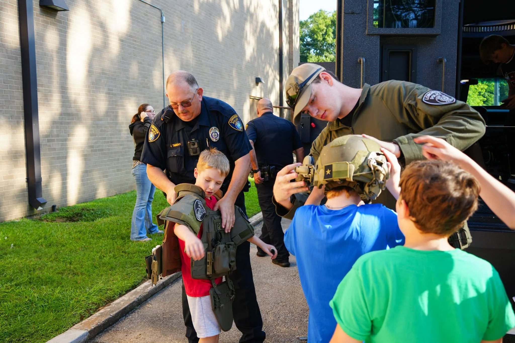 Children fitting protective military vests on at a community event with police and military personnel.
