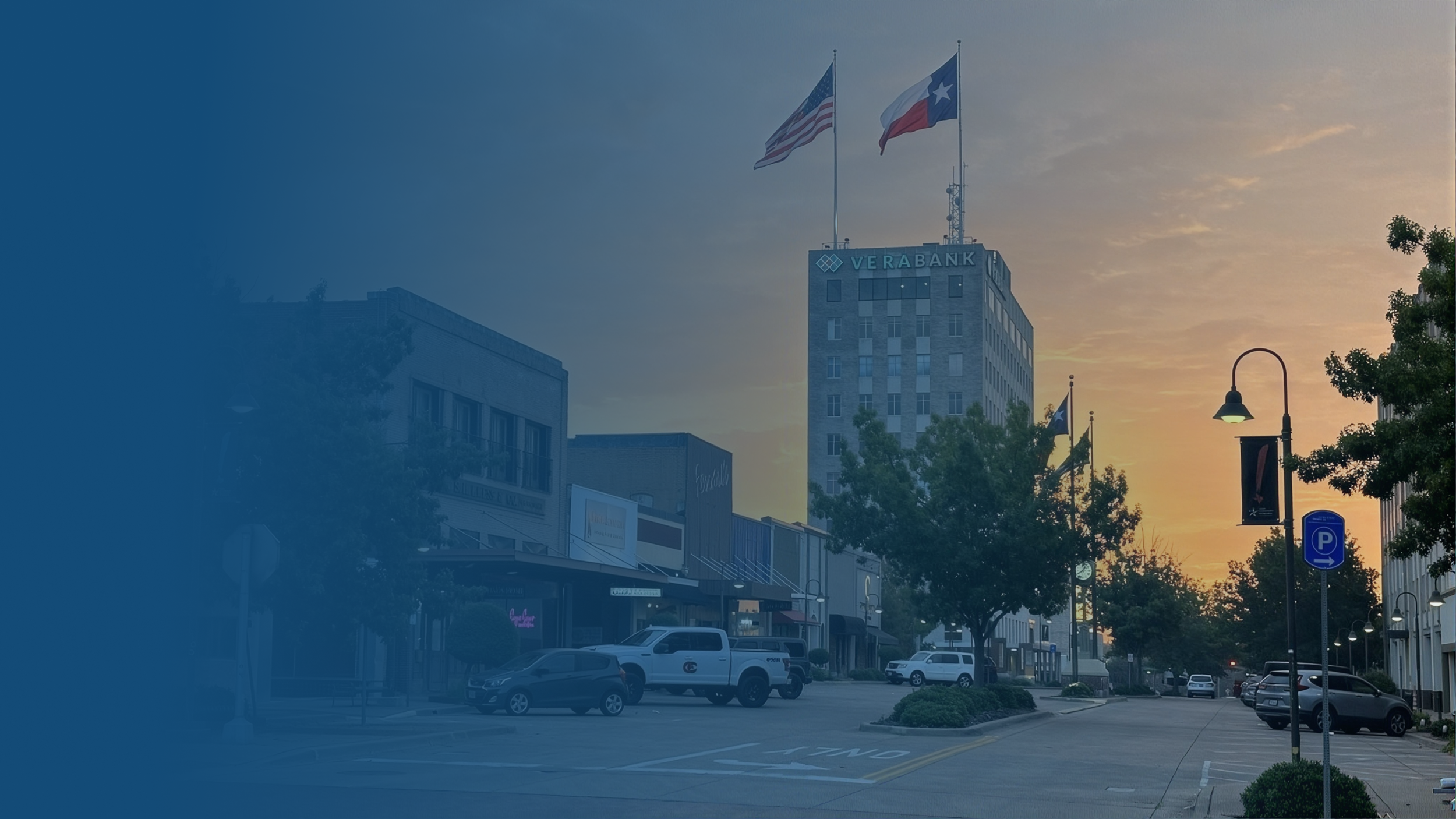 A city street with cars parked along the curb, a tall building with flags on top, trees lining the sidewalk, and a sunset sky in the background.