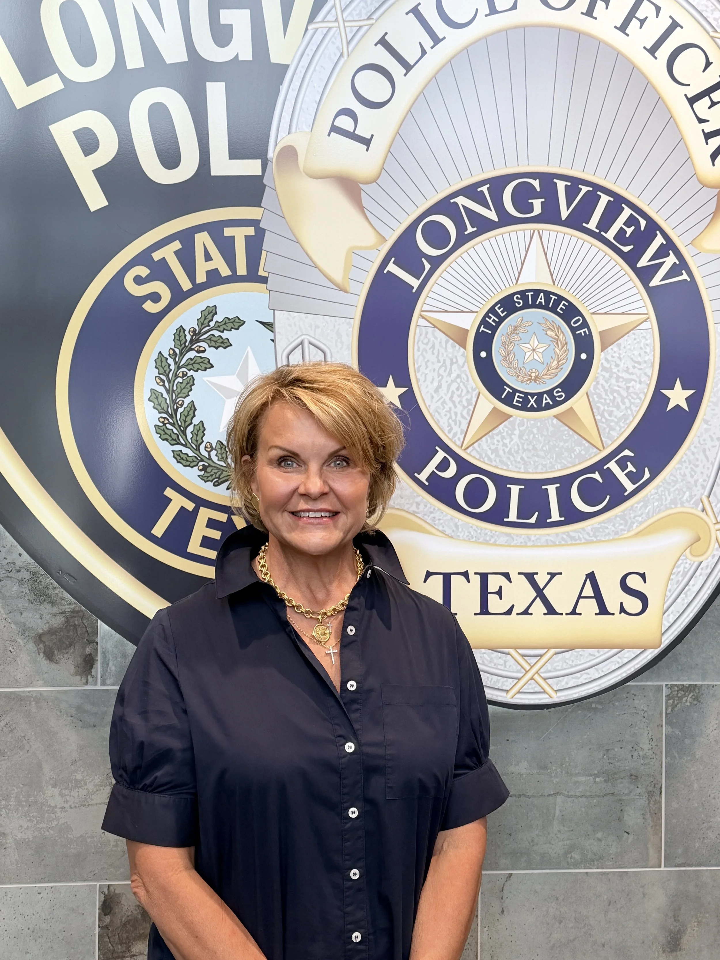 A smiling woman with short blonde hair, wearing a black shirt with pearl buttons and layered necklaces including a gold chain, a medallion, and a cross pendant, standing in front of a large police department emblem for the Longview Police Department in Texas.