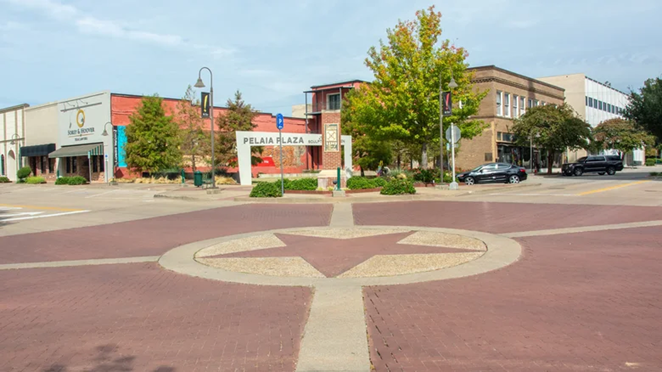 A small downtown area with a plaza in the center. There is a large arrow and circle design on the brick sidewalk. Surrounding buildings include retail shops and offices, with trees and streetlights. A sign reads 'Pelaia Plaza' with additional text that is not fully visible.