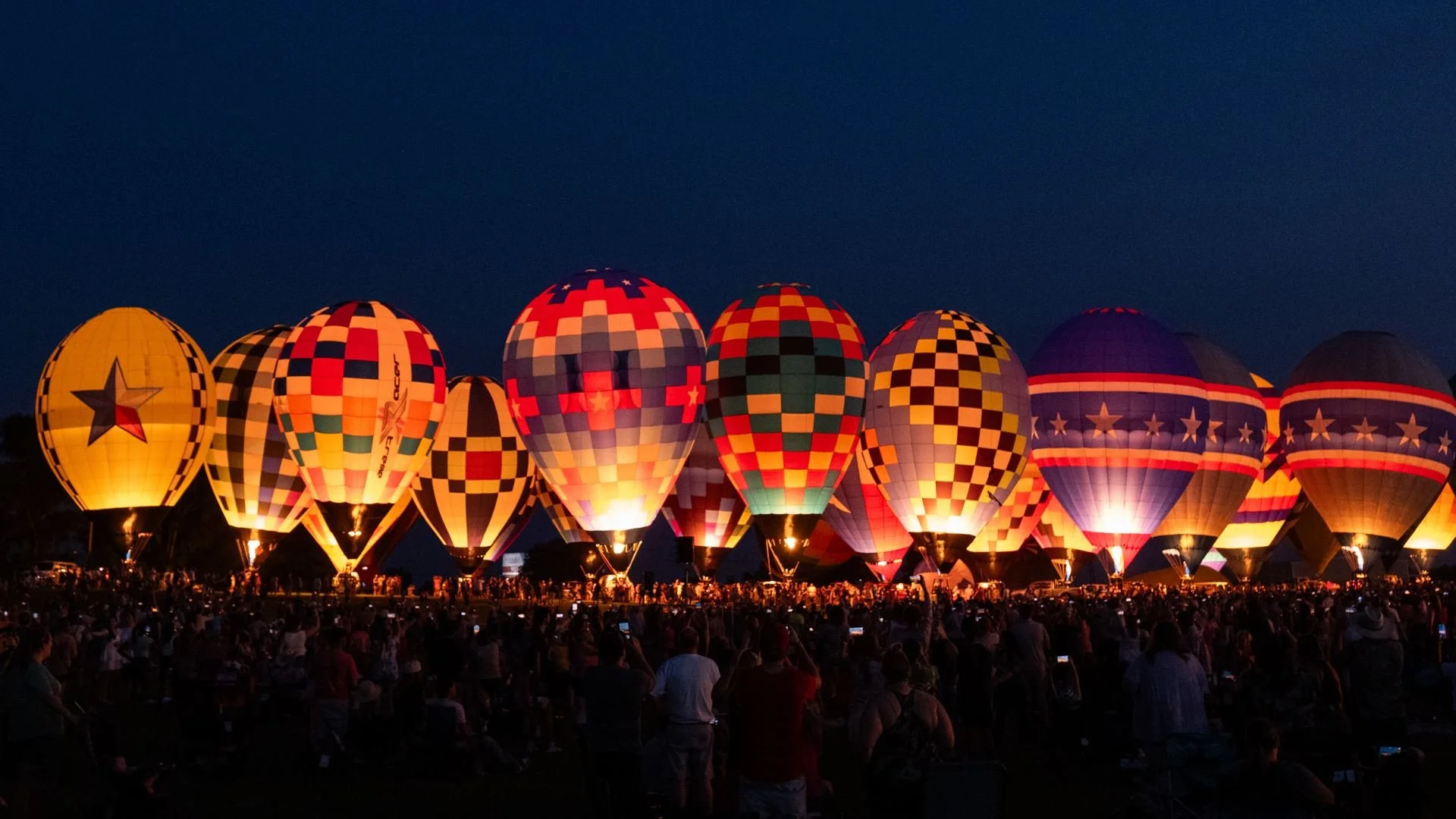 Nighttime scene of numerous illuminated hot air balloons with geometric patterns ready for ascent, gathered in a field with a large crowd of spectators.