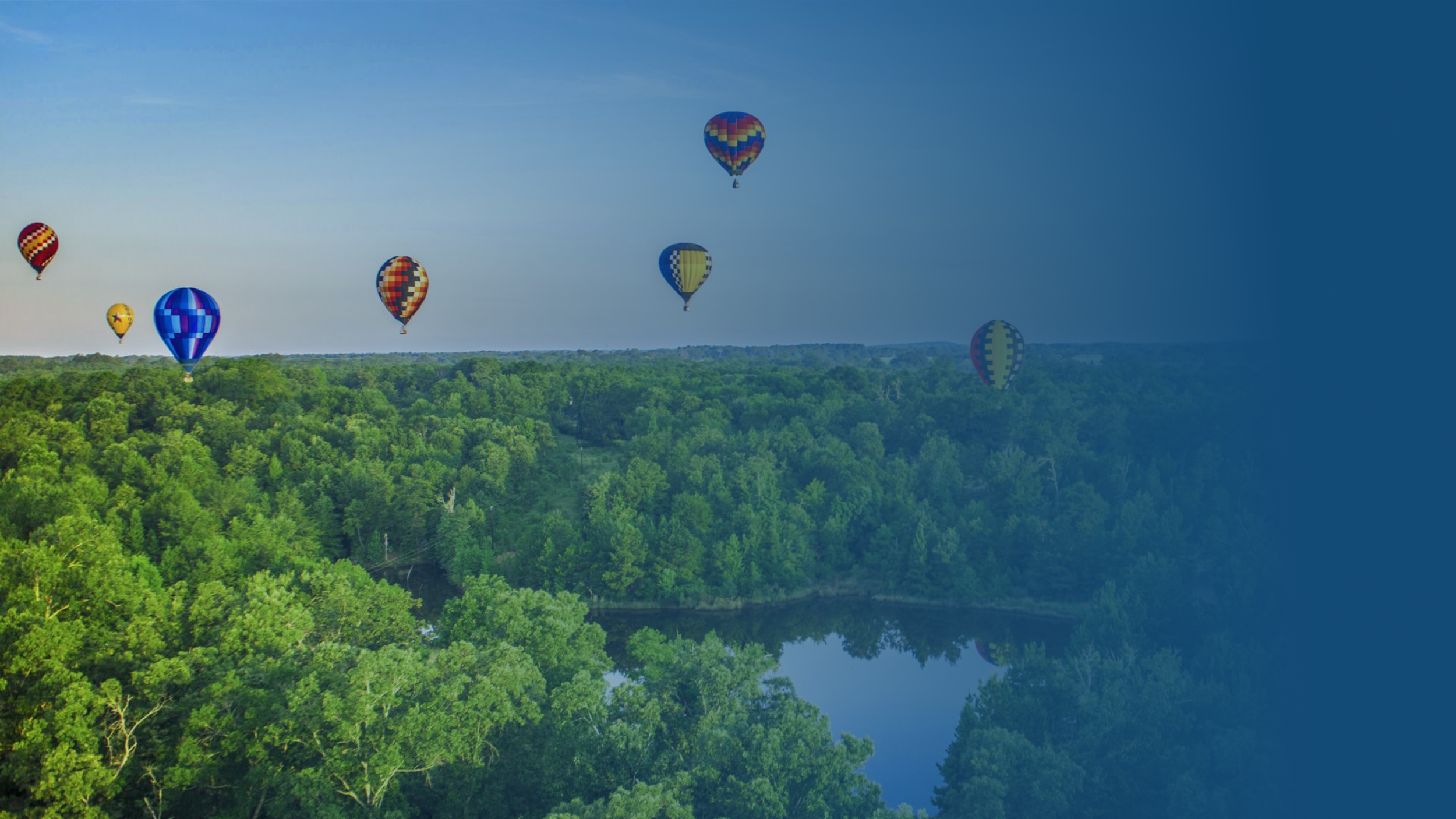 Multiple colorful hot air balloons floating above a lush green forest and a water body during daytime.