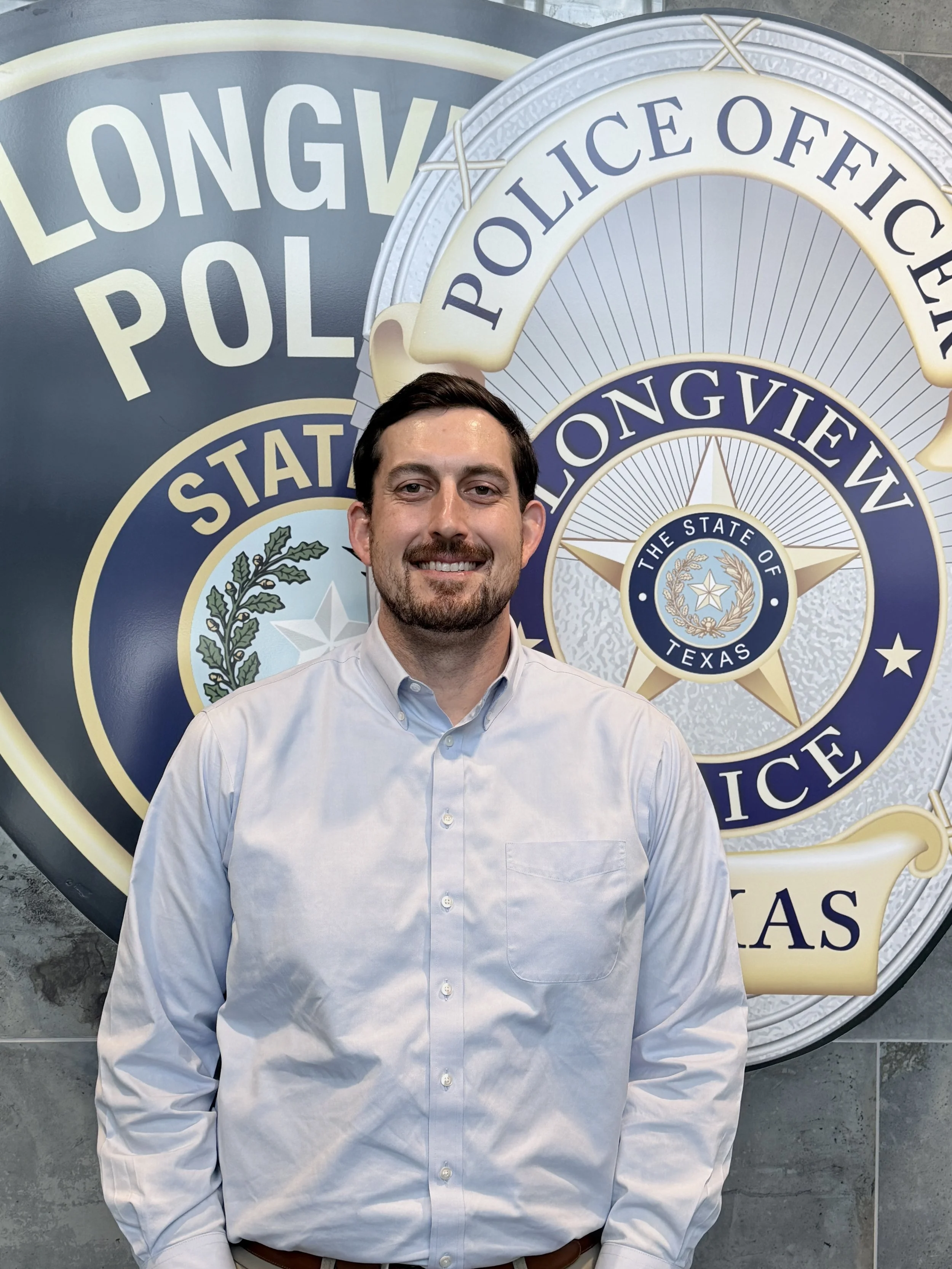 A smiling man in a light blue shirt standing in front of a Texas Longview Police sign.