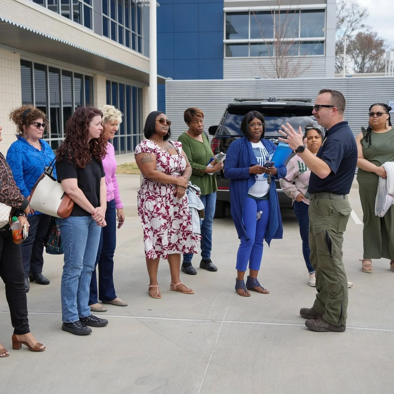 A man wearing glasses and a black shirt is speaking to a group of people outside a modern building. The group, mostly women, are listening attentively, some holding notebooks, phones, or bottles. The setting is an urban area with a car and a building with glass windows in the background, and the weather appears to be overcast.