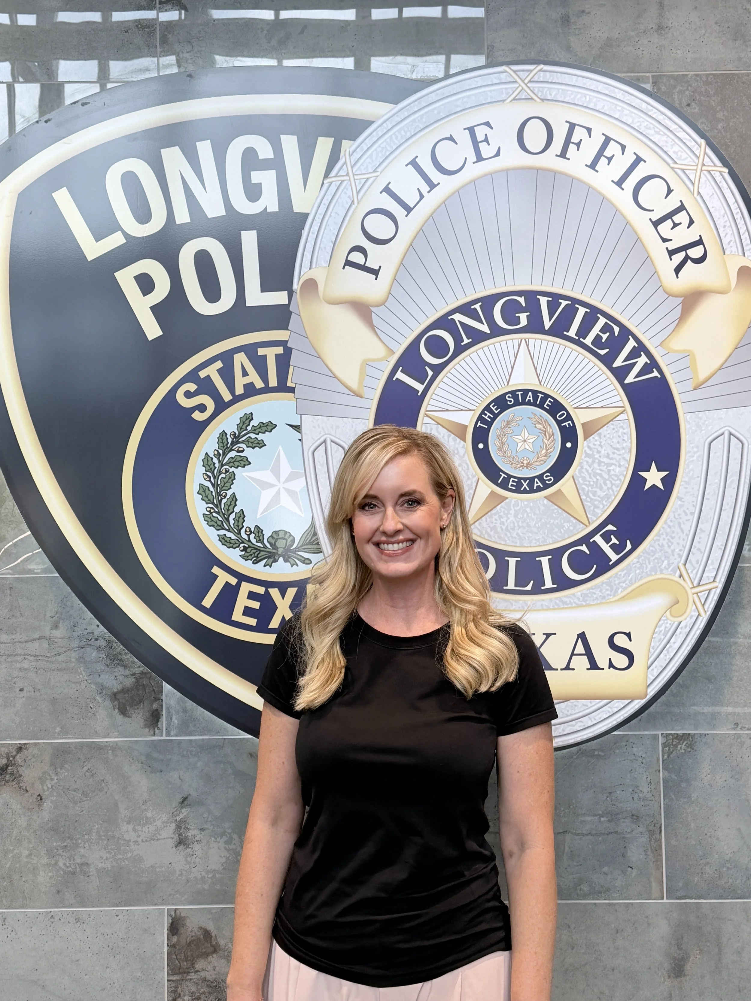 A woman with long blonde hair and a black shirt standing in front of a Texas police badge display.