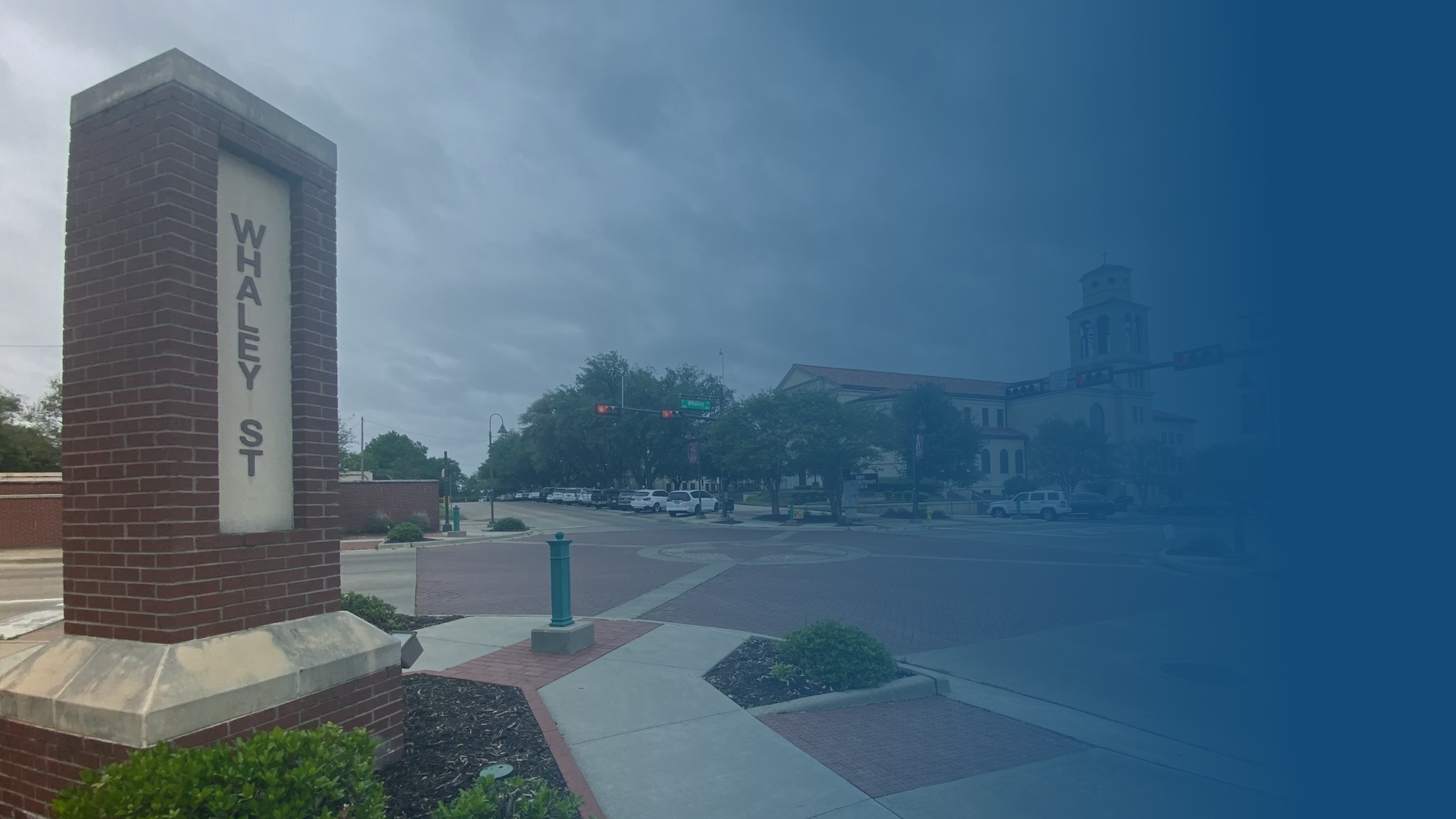 Street view of Whaley Street with a brick sign pillar, parked cars, trees, and a building with a tower in the background under a cloudy sky.