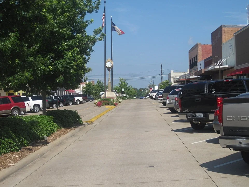 A small-town street with parked cars on each side, trees, a clock tower displaying the time as 2:20, and flags of the United States and France flying on poles.