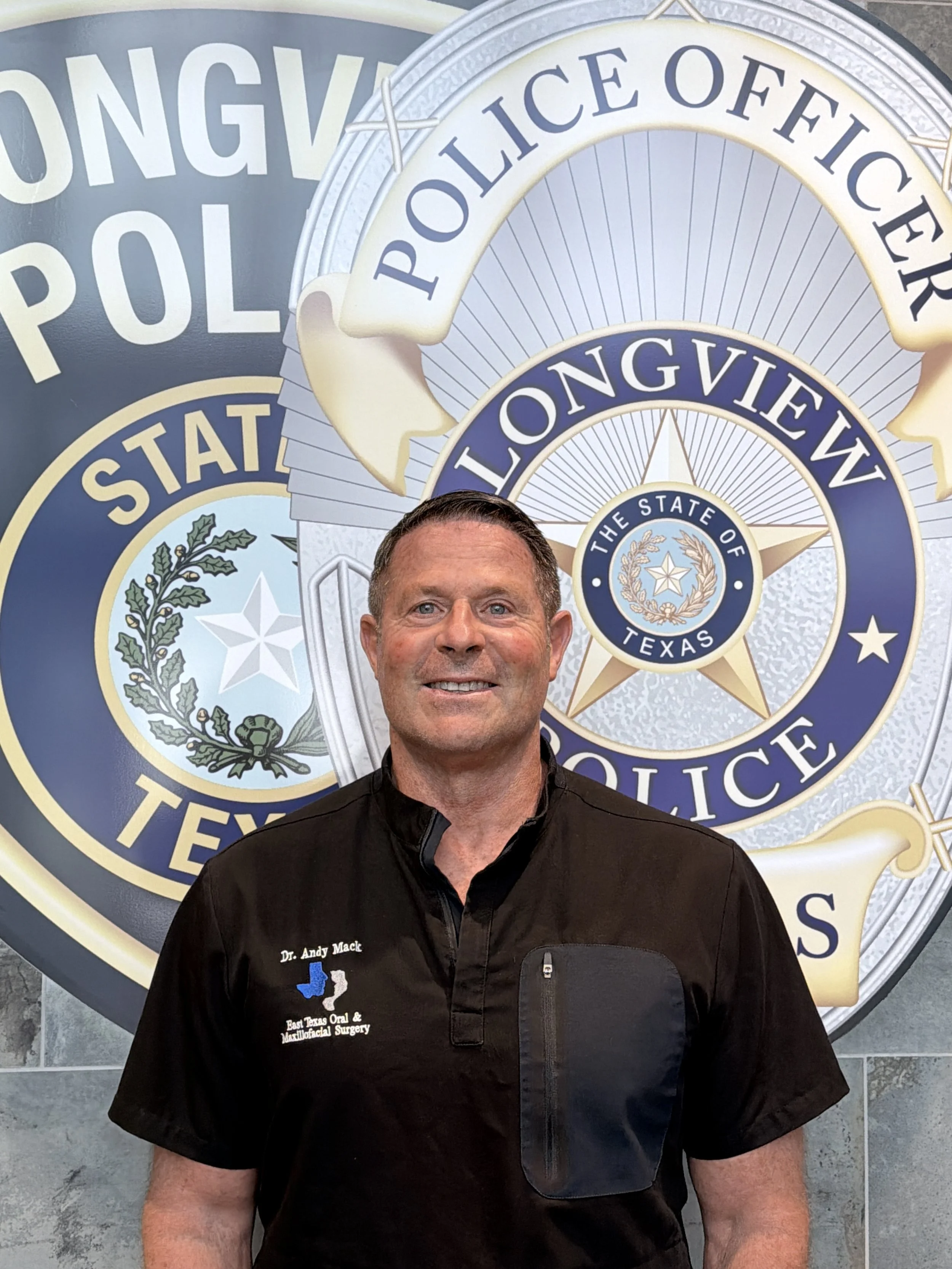 A man wearing a black medical uniform standing in front of a Texas police badge display. The uniform has a logo that says East Texas Oral & Maxillofacial Surgery.