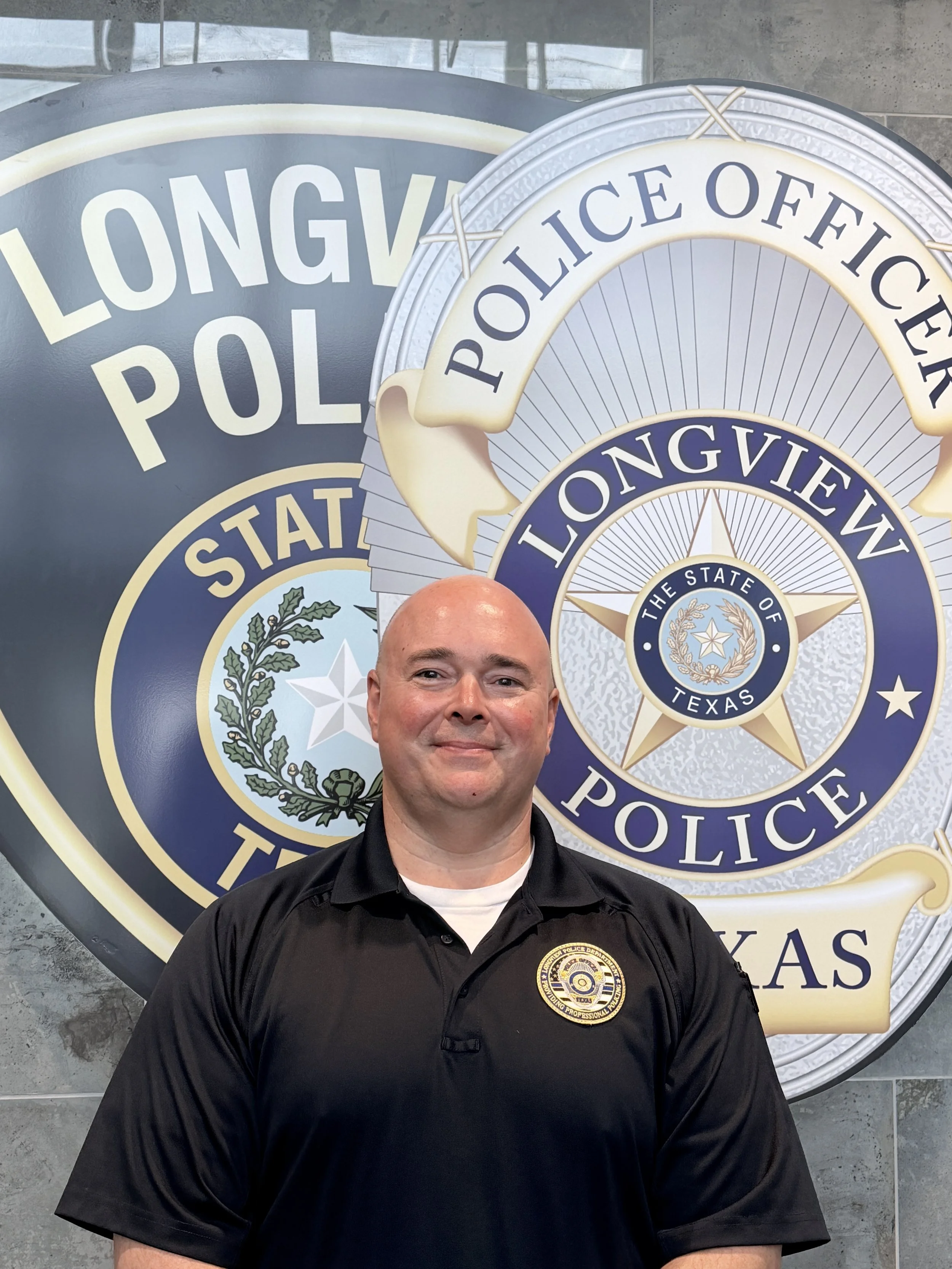 A police officer wearing a black uniform and badge stands in front of large police department emblems, including the Longview Police badge, at a police station.