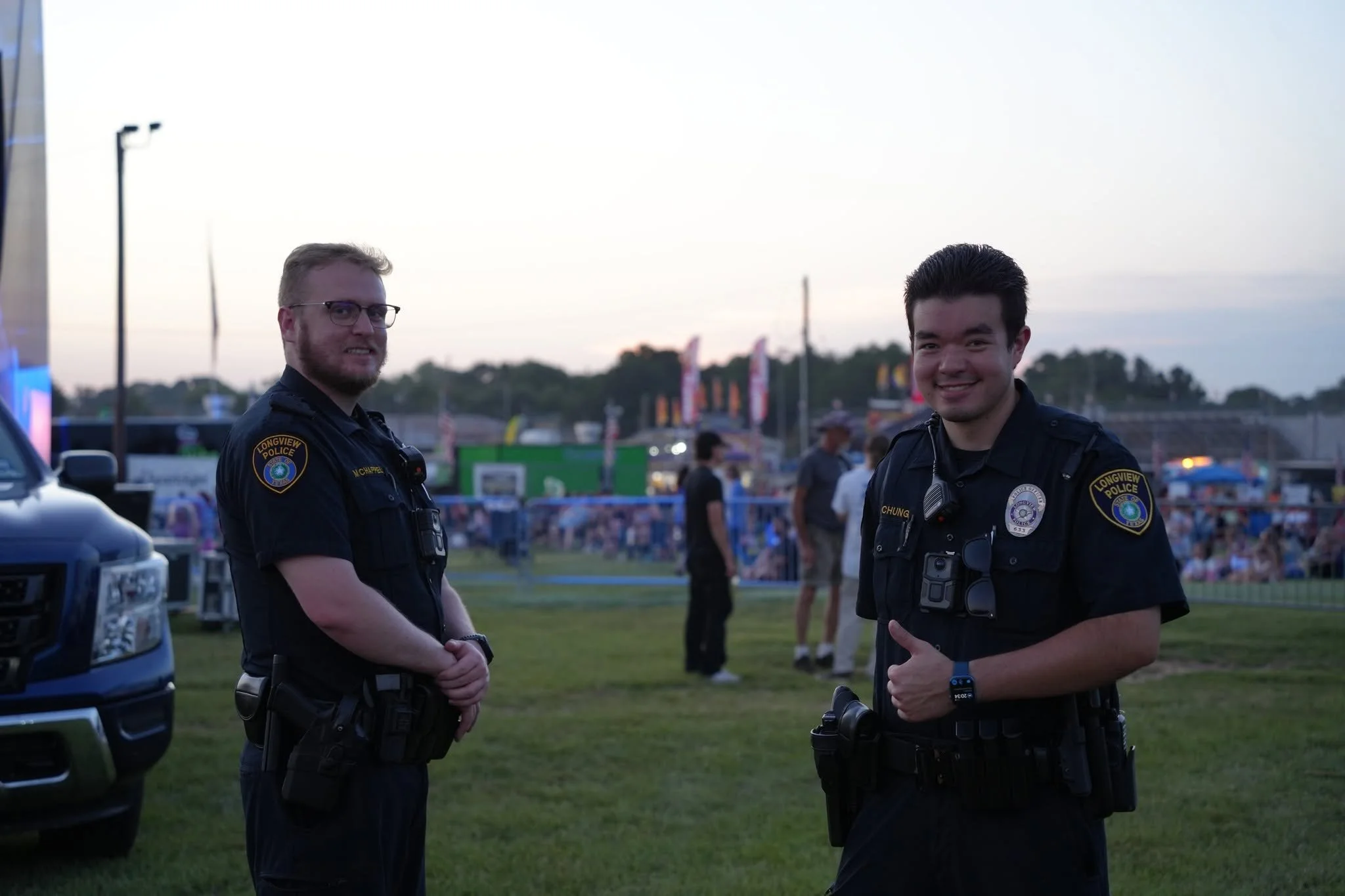 Two police officers, one male and one female, stand outdoors at dusk. They are facing the camera, smiling, with a crowd and a fairground in the background.
