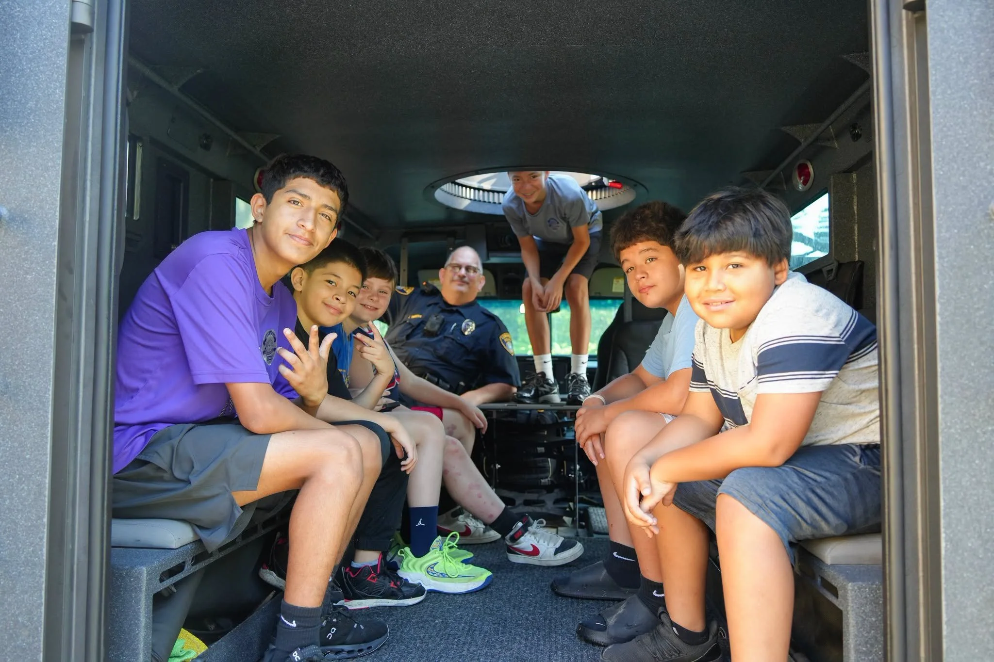 A group of boys and a police officer sitting inside a police vehicle, with two boys and an officer visible through a hatch in the vehicle ceiling, smiling and looking at the camera.