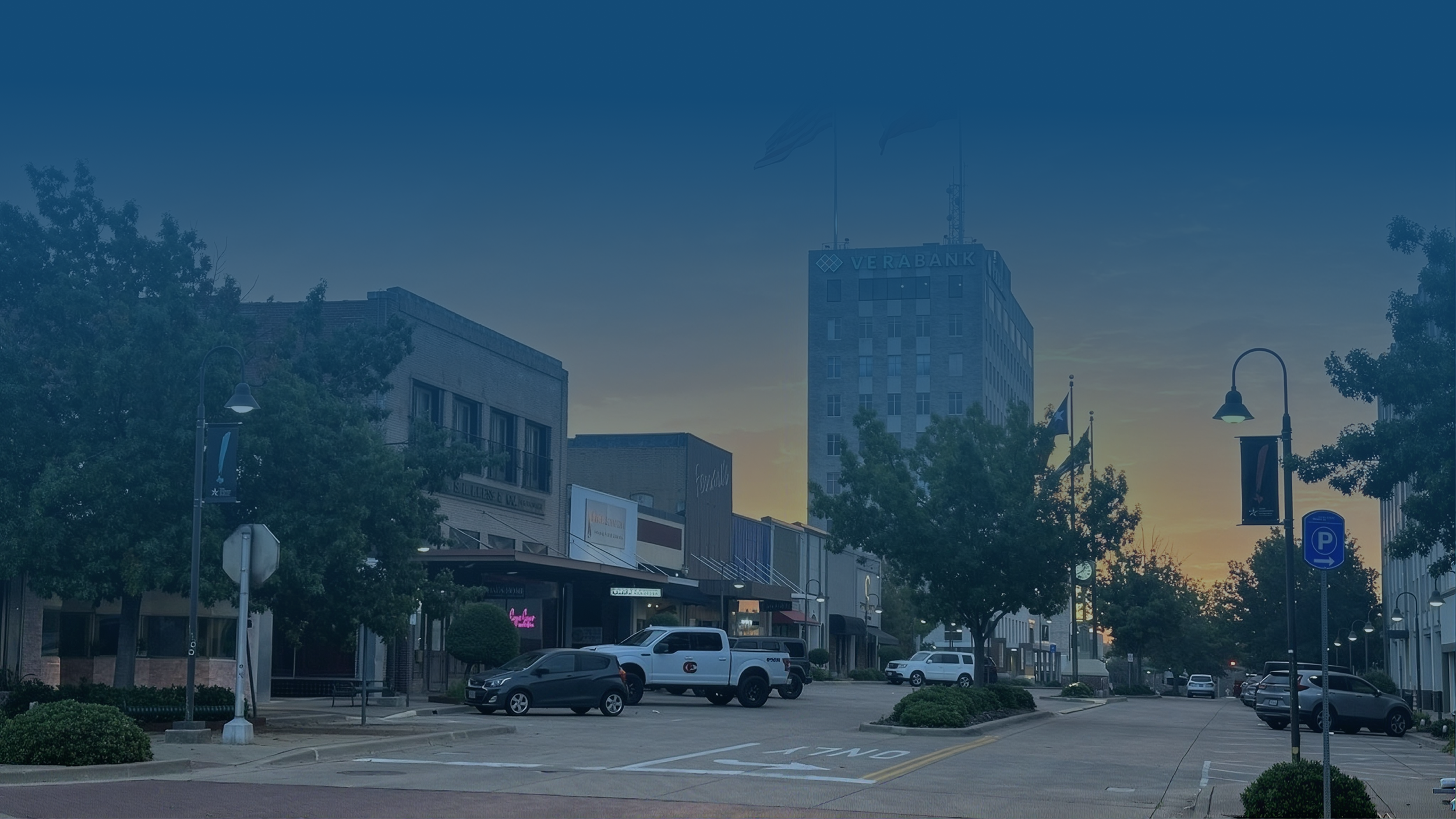 A city street at dusk with buildings, parked cars, trees, street lamps, and flags in the background.