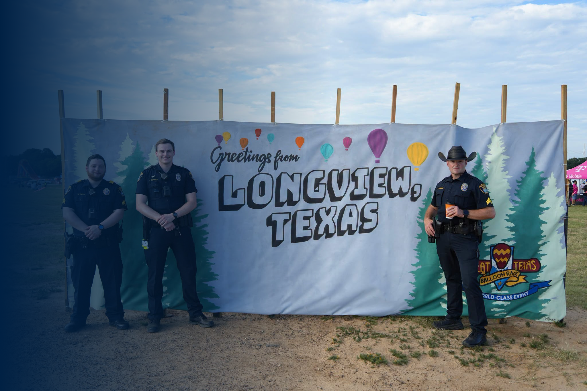 Three police officers standing in front of a colorful banner that reads "Greetings from Longview Texas," with trees and a partly cloudy sky in the background.