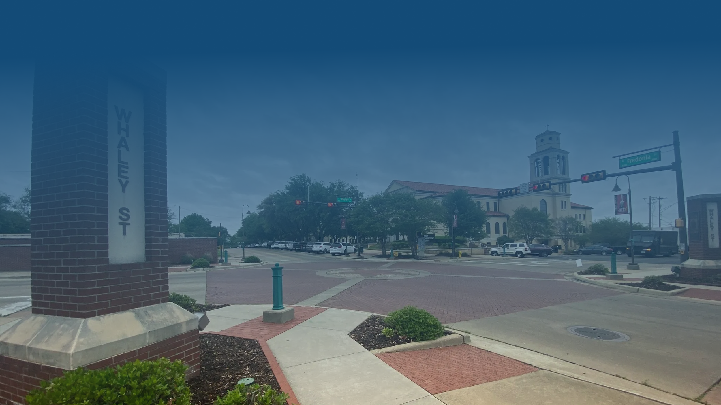 View of the town square with a brick pillar showing 'Whaley St', a street sign for Fredonia Ave, a church building with a bell tower, parked cars, and a bus at a traffic light under a cloudy sky.