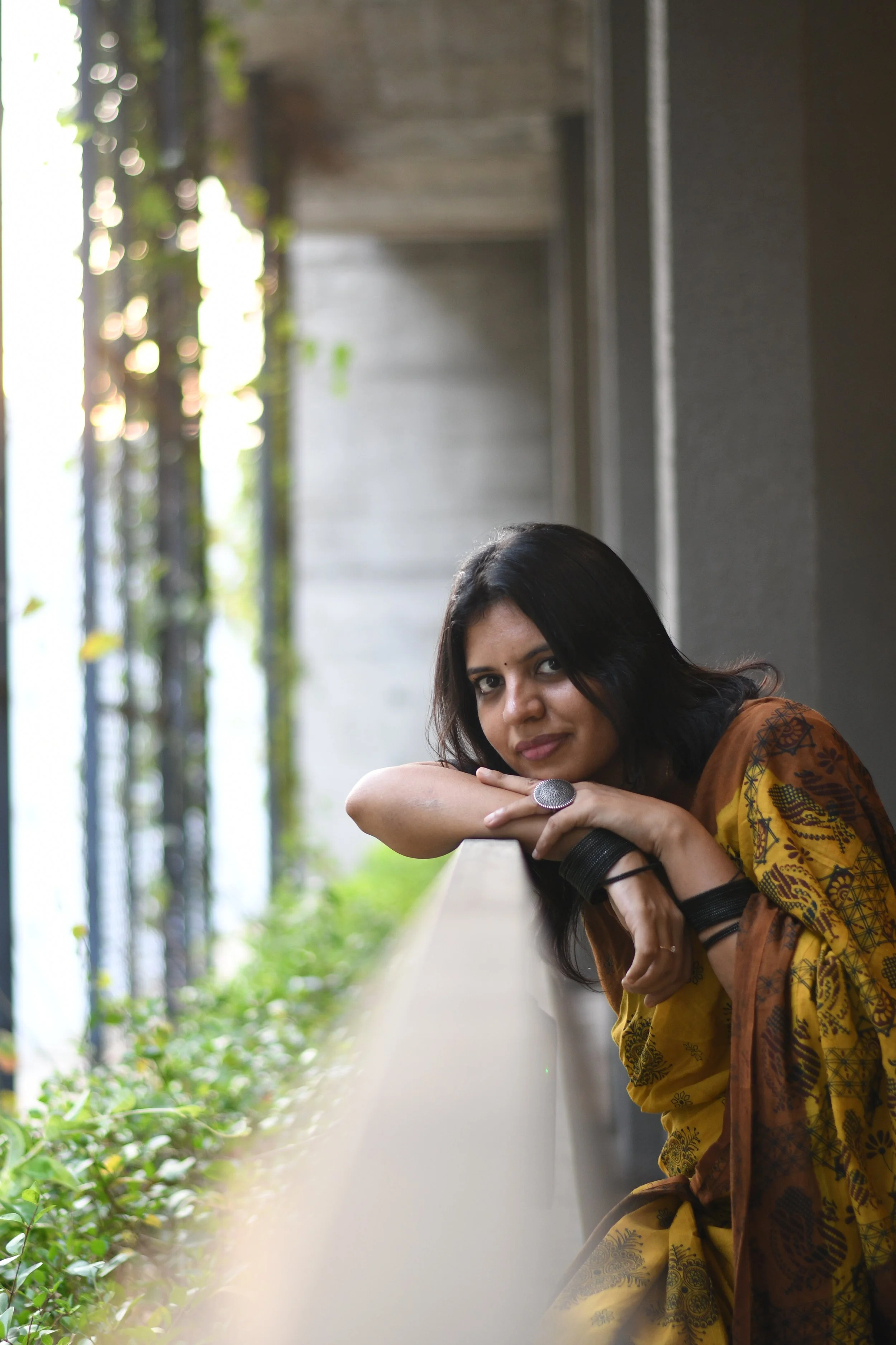 R.M.Kritika, Author of 'Dear Sh'. A book about hope, survival, coming of age, literary fiction. Contemporary wearing a yellow and brown traditional dress with black bangles, leaning on a railing with plants on the side and sunlight in the background.