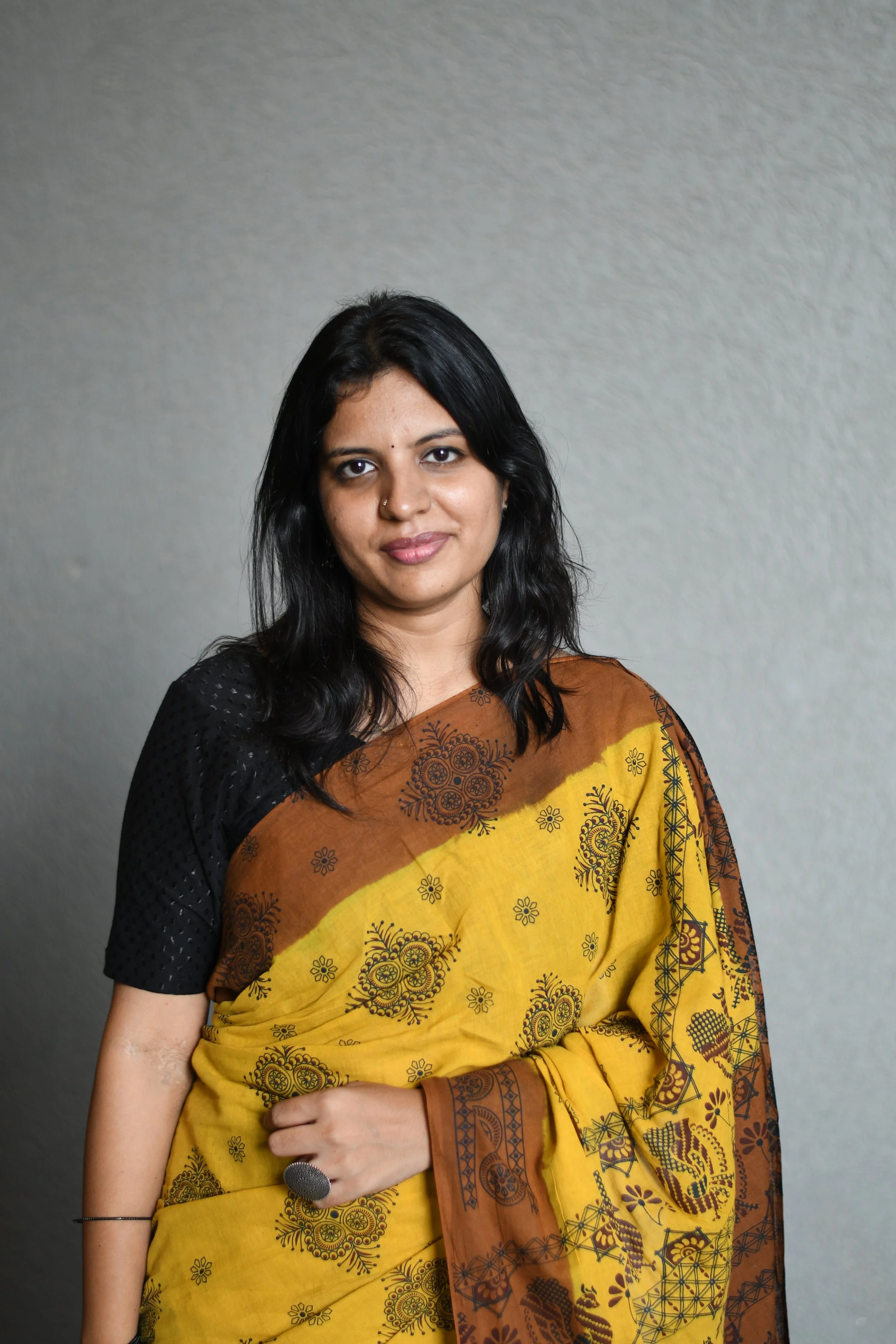 A woman with black hair and a nose ring dressed in a yellow and brown patterned sari, standing against a gray wall.