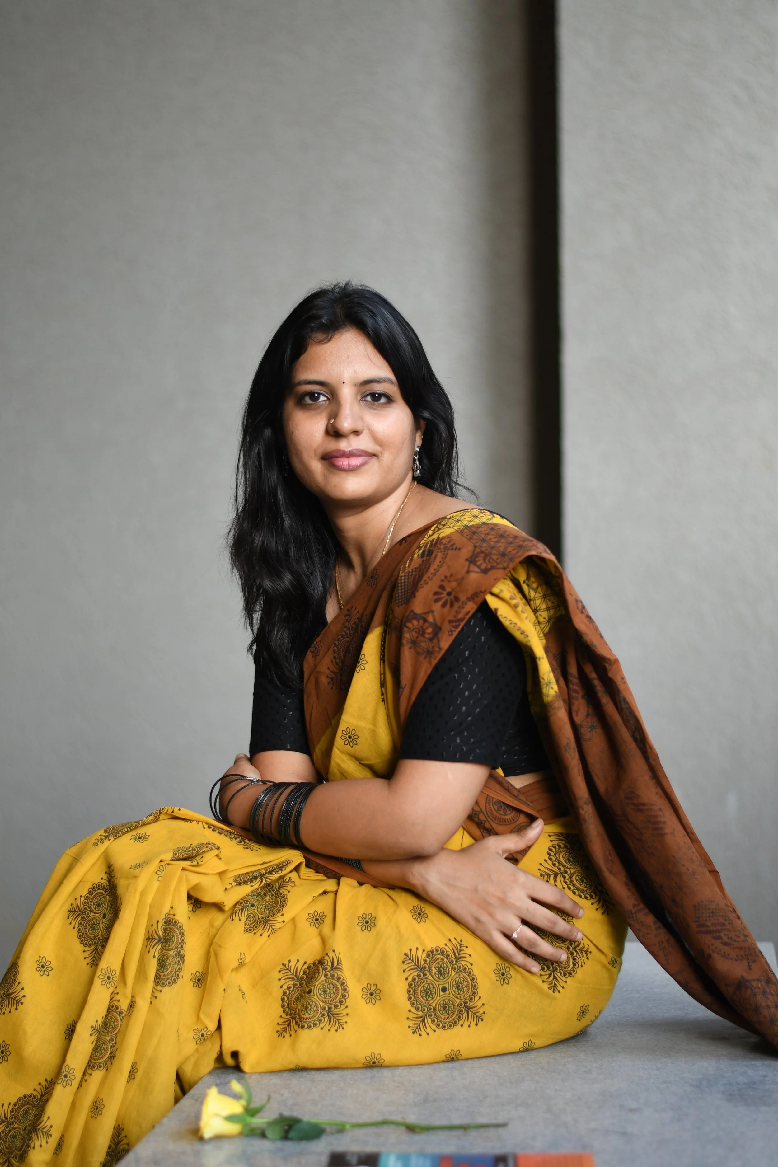 R.M.Kritika, Author of 'Dear Sh'. Sitting on the floor, wearing a traditional Indian saree in black and yellow. She has long black hair, a nose ring, and is looking at the camera. There is a yellow rose on the floor in front of her.