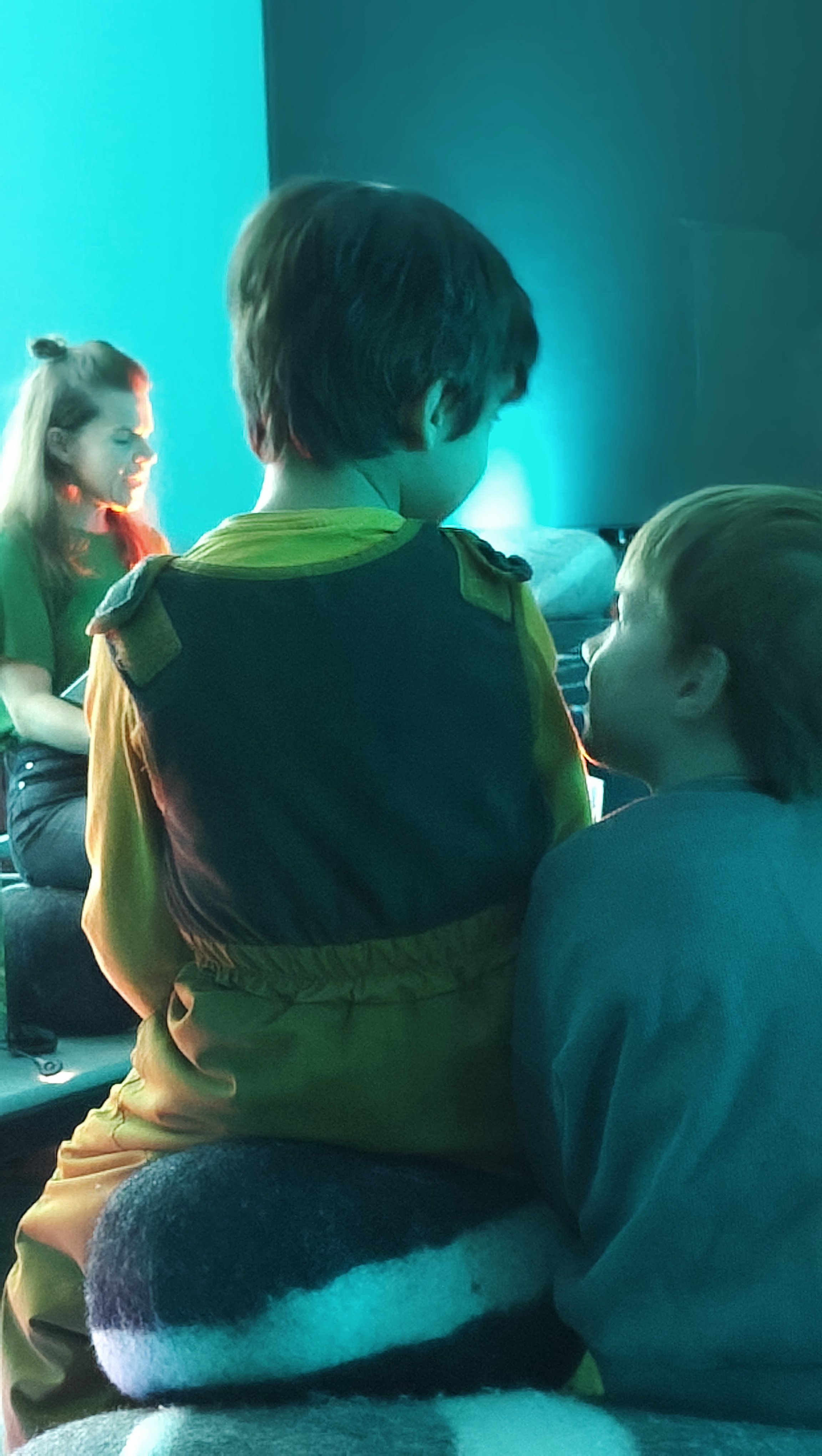 Drei Kinder sitzen in einem dunklen Raum mit blauer Beleuchtung und unterhalten sich. Zwei Kinder sind sichtbar, eines hat braune Haare und trägt ein grünes Hemd, das andere hat blonde Haare und trägt einen blauen Pullover.
