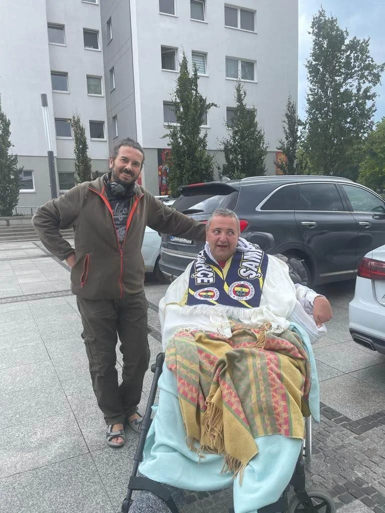 Zwei Männer im Freien, einer sitzt im Rollstuhl mit Decken und Schal, der andere steht neben ihm und berührt ihn freundlich auf die Schulter, im Hintergrund stehen Autos und ein modernes Wohngebäude.