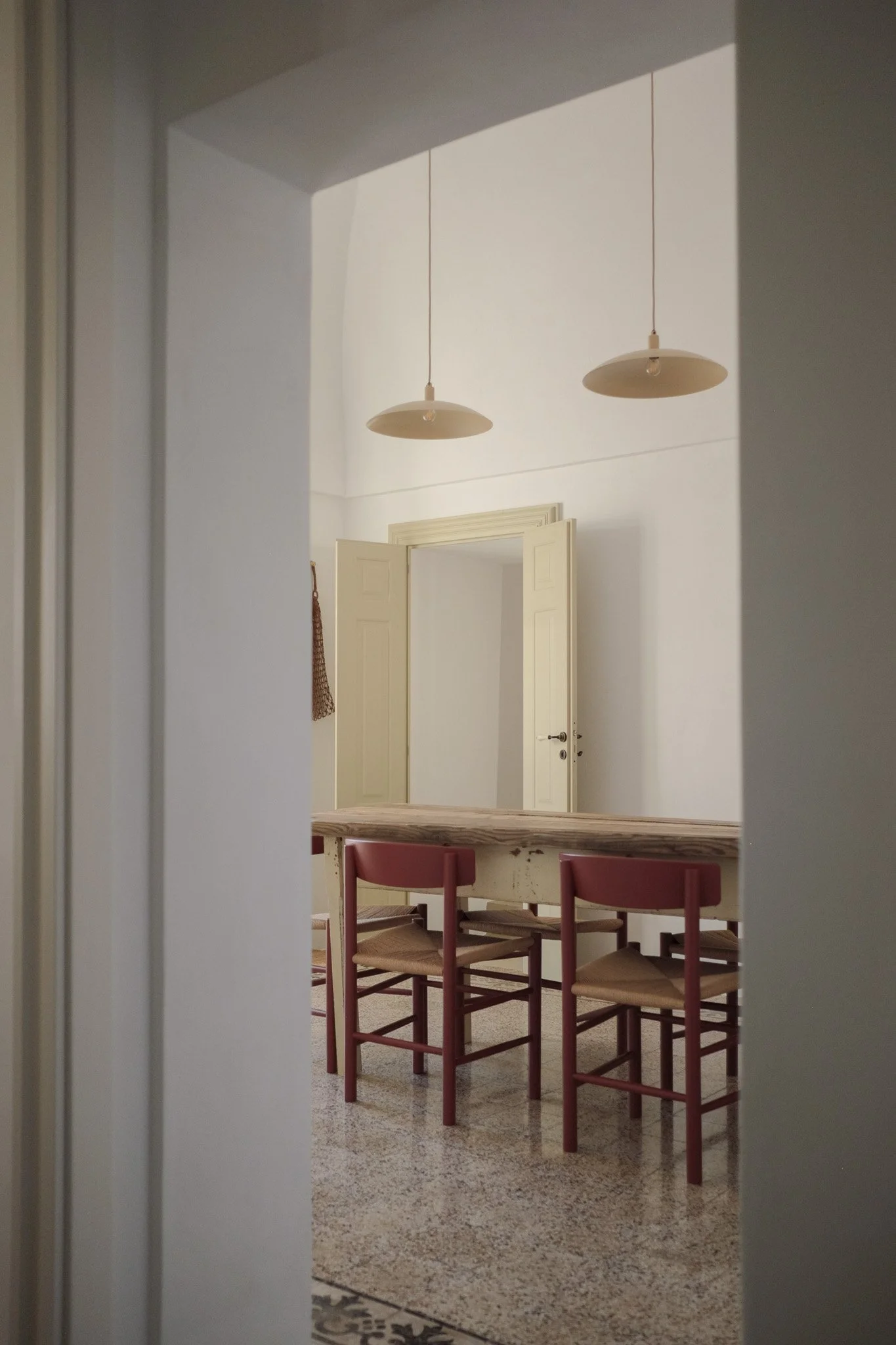 View through a doorway showing a dining area with a long table and multiple chairs, white walls, two hanging ceiling lamps, and a door in the background.