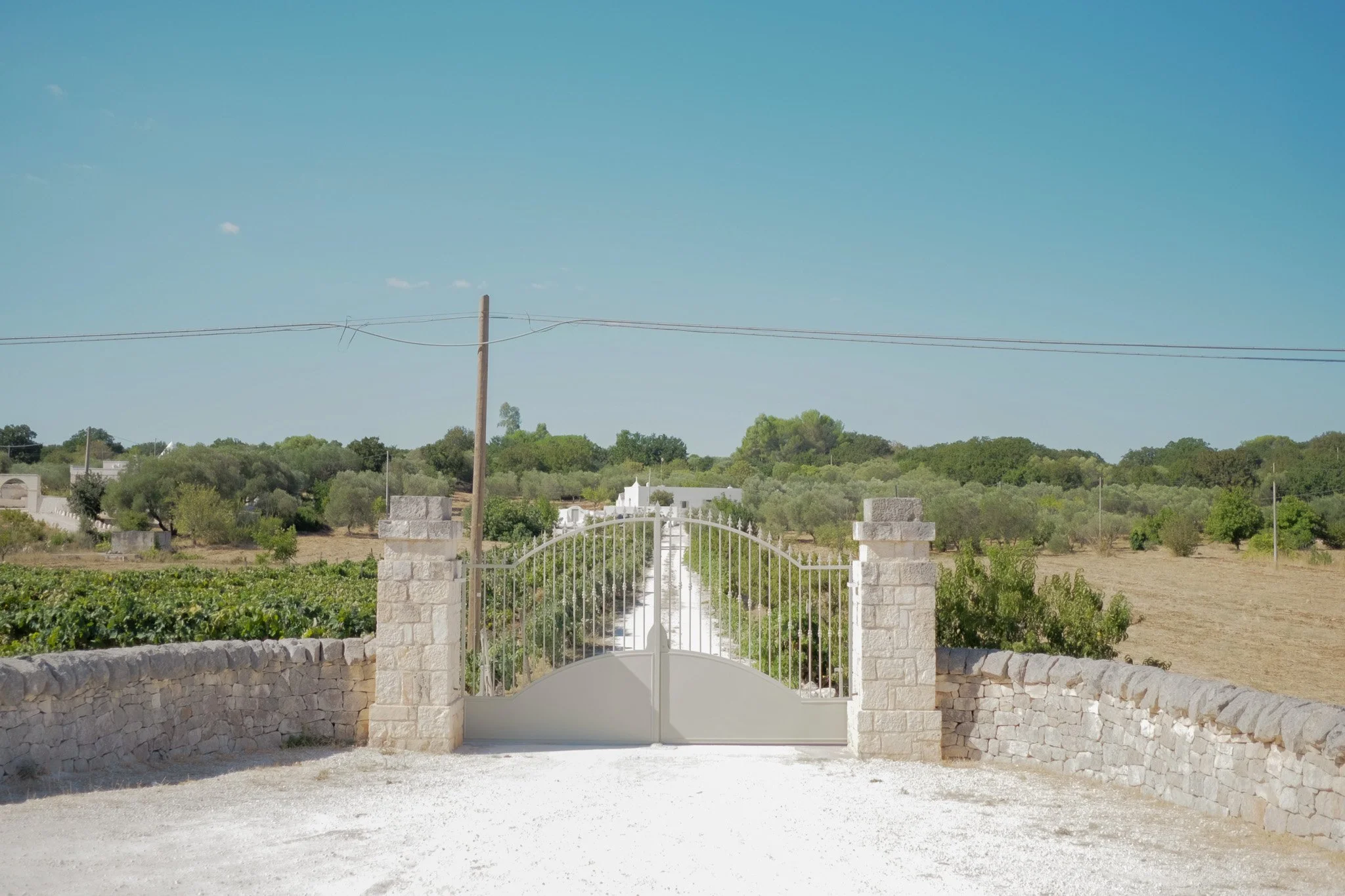 White gate with stone pillars leading into a rural landscape with trees, fields, and a clear blue sky.