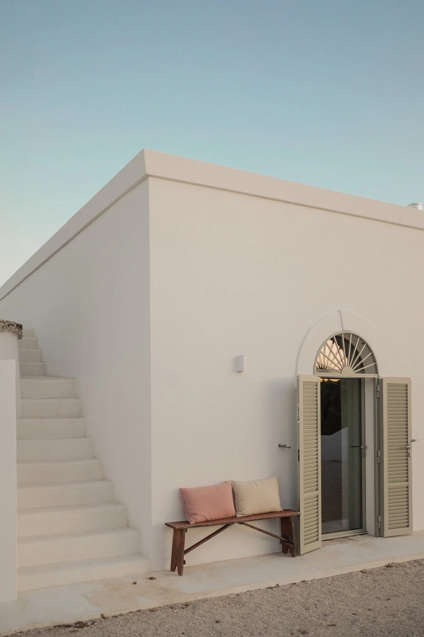 Exterior of a white building with stairs, an arched window with shutters, and a wooden bench with pink and beige pillows.