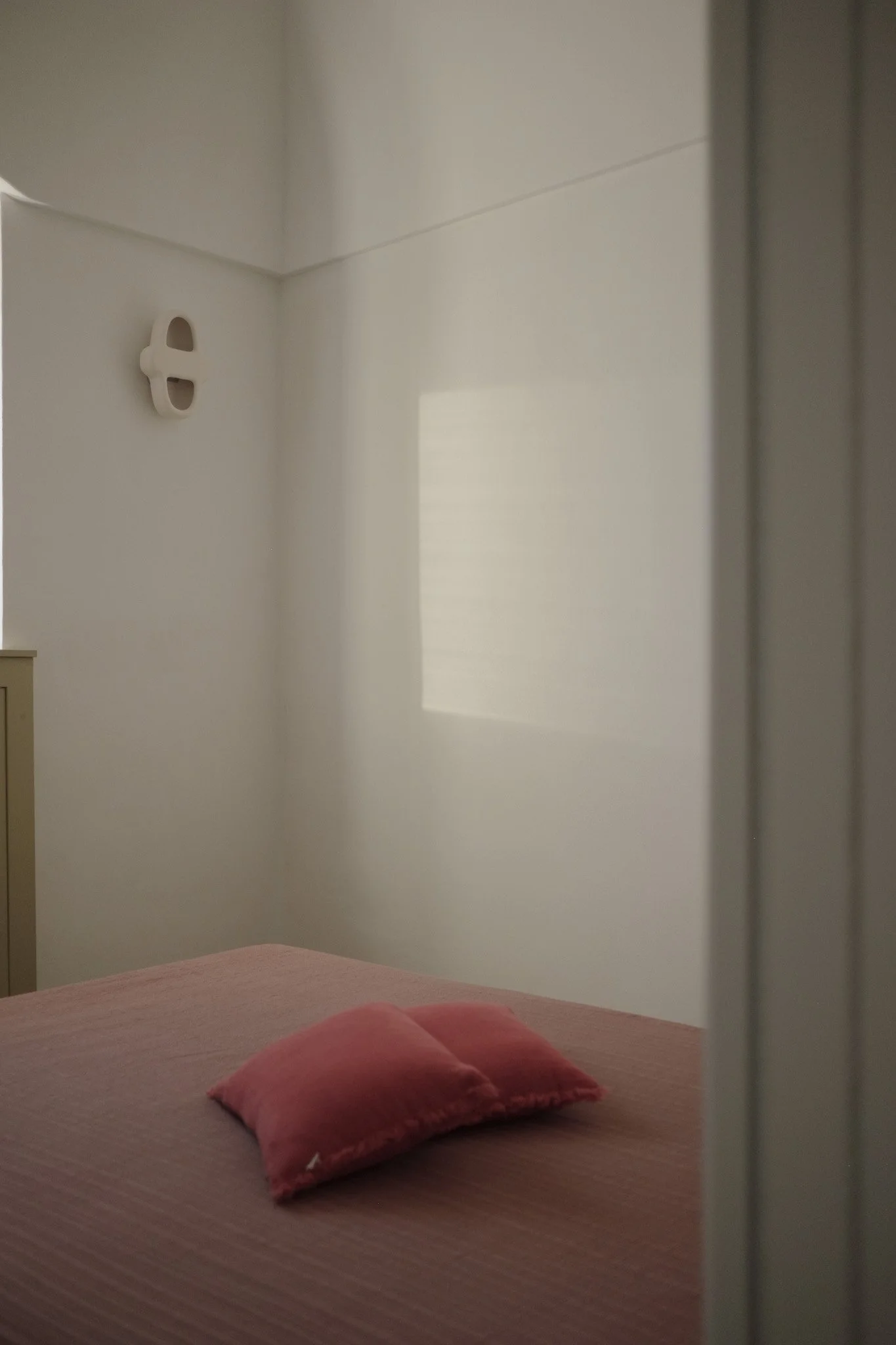 View of a bedroom with a bed covered in a pink bedspread and two pink pillows. The wall is white with a window that has a shade or curtain, and a white wall fixture is visible in the corner.