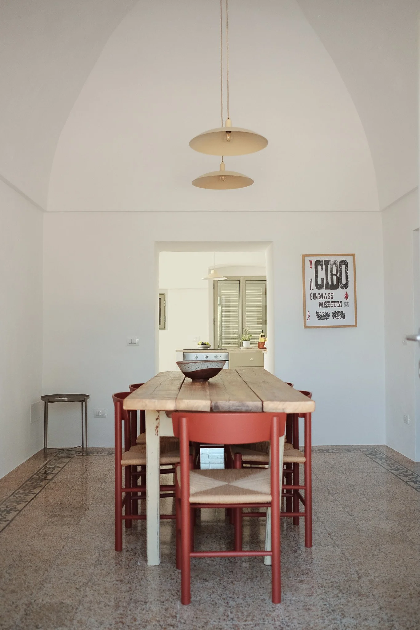 A dining room with a wooden table surrounded by six red chairs. The room has a tiled floor with a decorative border, white walls, and an arched ceiling. A framed poster hangs on the right wall. In the background, there is a kitchen area visible throu