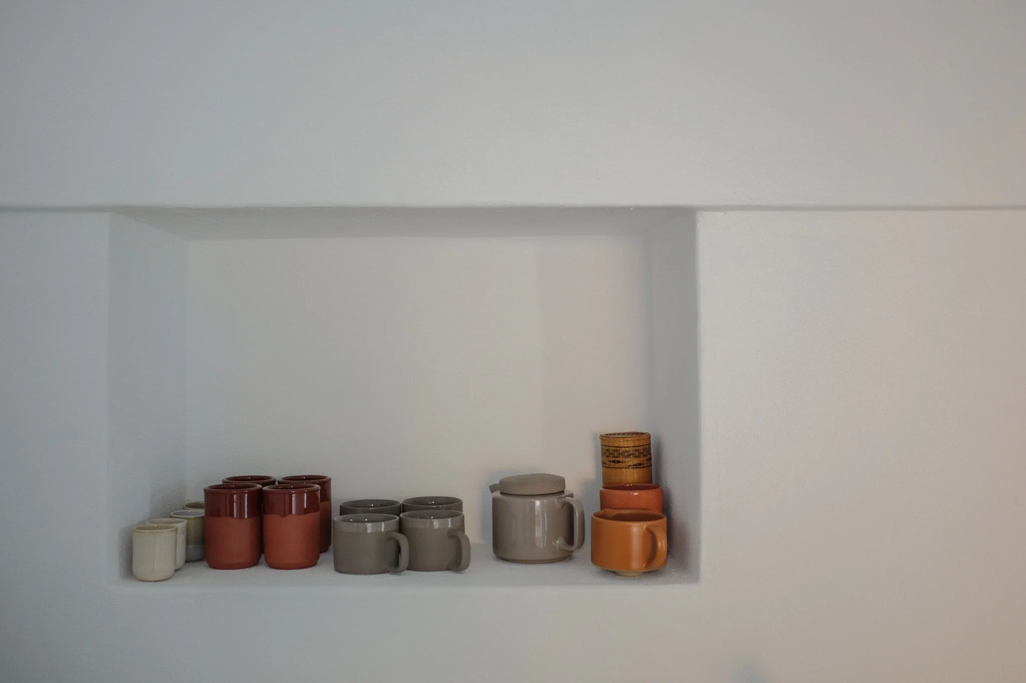 A white shelf holding various ceramic mugs and cups in earthy tones, with a small jar and a wooden container on the right side.