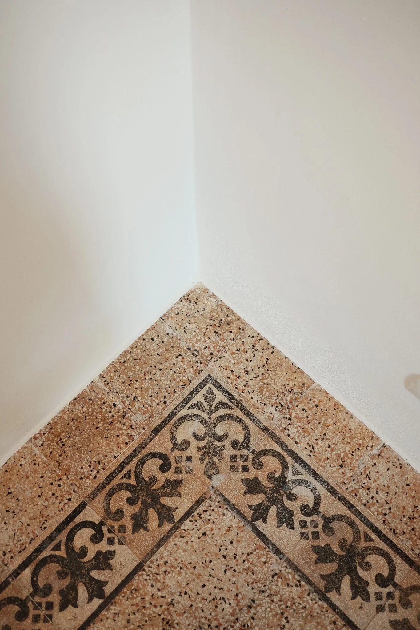 Corner of a tiled floor and white walls, with an ornate black decorative pattern on the tile near the bottom of the image.